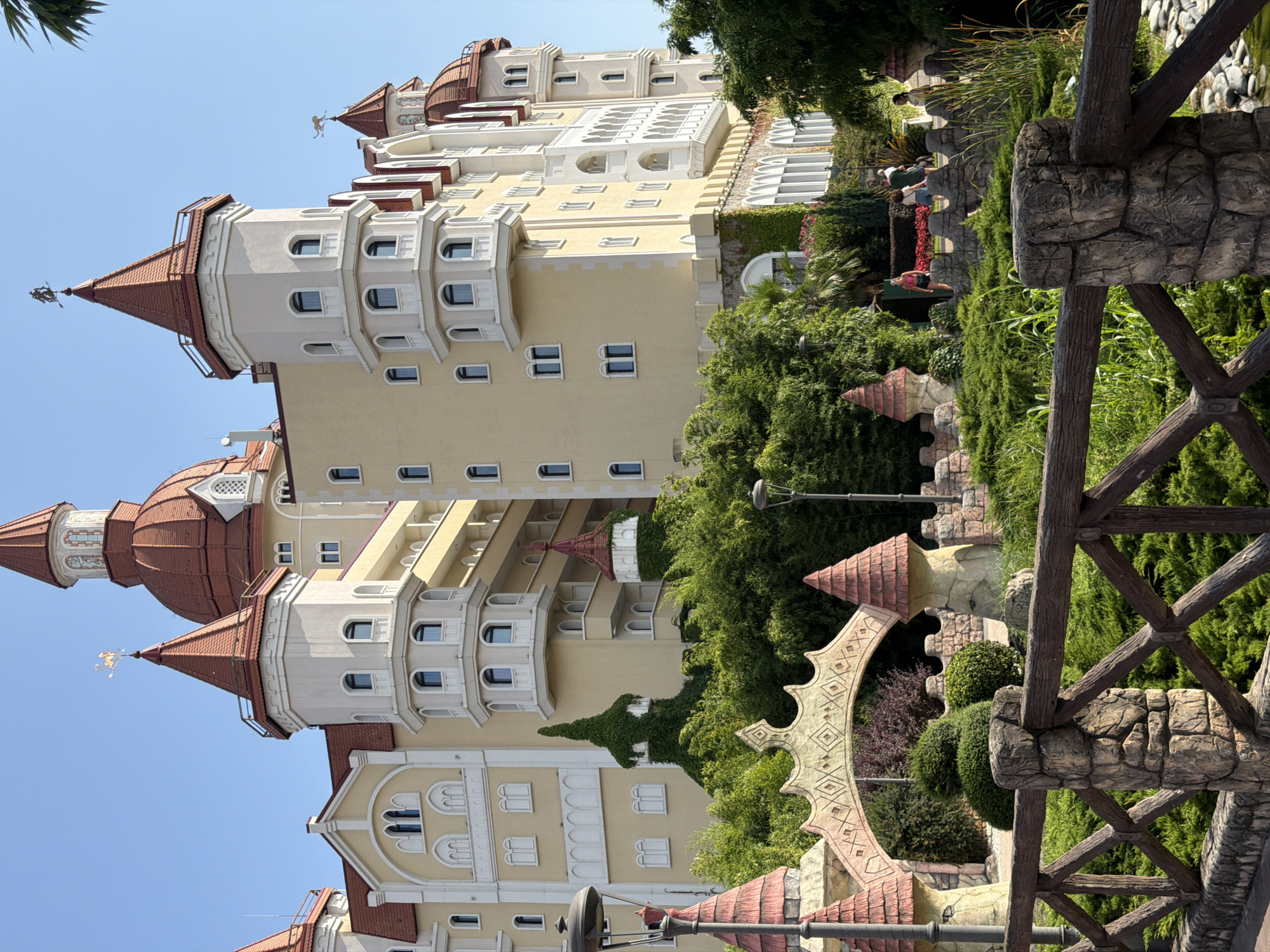 In the city of Sirius, Russia, a large, multi-story building with a fairy-tale architectural style is visible. The building features numerous turrets and domes, primarily colored in red and yellow, with white decorative elements. The structure is set against a clear blue sky. In the foreground, there is a lush green garden with various plants and trees. A wooden fence with stone posts separates the foreground from the garden. Several small, decorative structures resembling turrets with red roofs are present within the garden. A decorative archway with an intricate pattern is also visible in the garden. A few individuals are seen walking through the garden in the middle ground. The overall scene depicts a well-maintained, whimsical outdoor environment adjacent to an elaborate building.