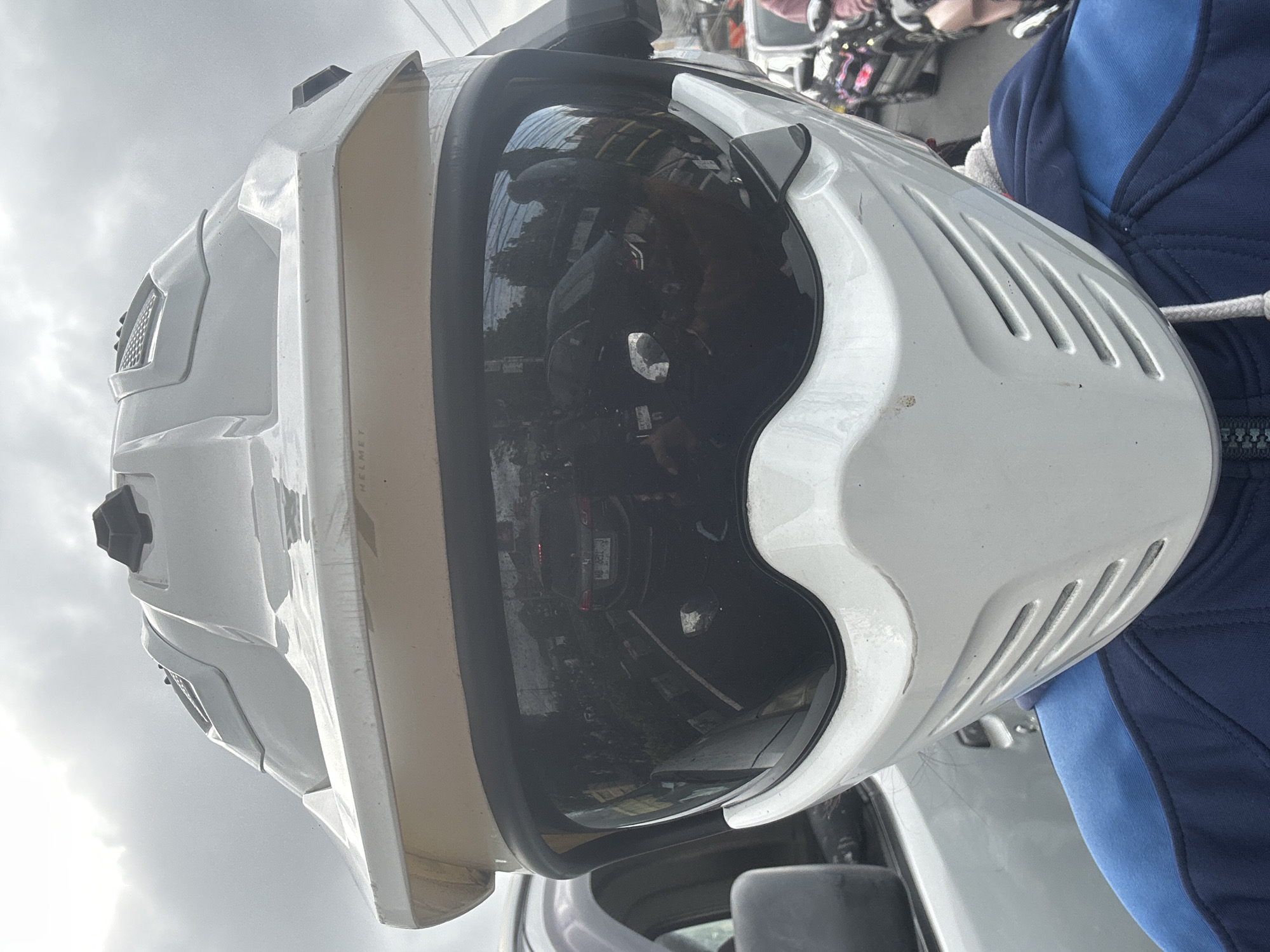 This is a close-up, vertically oriented photograph of a white motocross helmet. The helmet is the primary subject, filling most of the frame. It has a prominent visor that reflects the surrounding environment, showing glimpses of vehicles, buildings, and possibly other people. The helmet has vents and a textured surface, suggesting it's designed for protective purposes.
To the right of the helmet, a portion of a person wearing a blue jacket is visible, providing a sense of scale and context. The jacket appears to be made of a fabric material, with white drawstring visible.
The background consists of a cloudy sky, indicating overcast weather. The reflection in the helmet's visor also suggests an outdoor setting, possibly a street or parking area, with the presence of vehicles and what looks like a street curb. The image appears to have been taken during the daytime, given the overall brightness.
A small amount of text is visible on the helmet itself, oriented vertically. The letters "HELMET" can be discerned, confirming the nature of the object.
The image is focused on the helmet, with the person and background elements serving as secondary context. There are no strong emotional cues conveyed in the image. The scene is set in Denpasar, Indonesia.