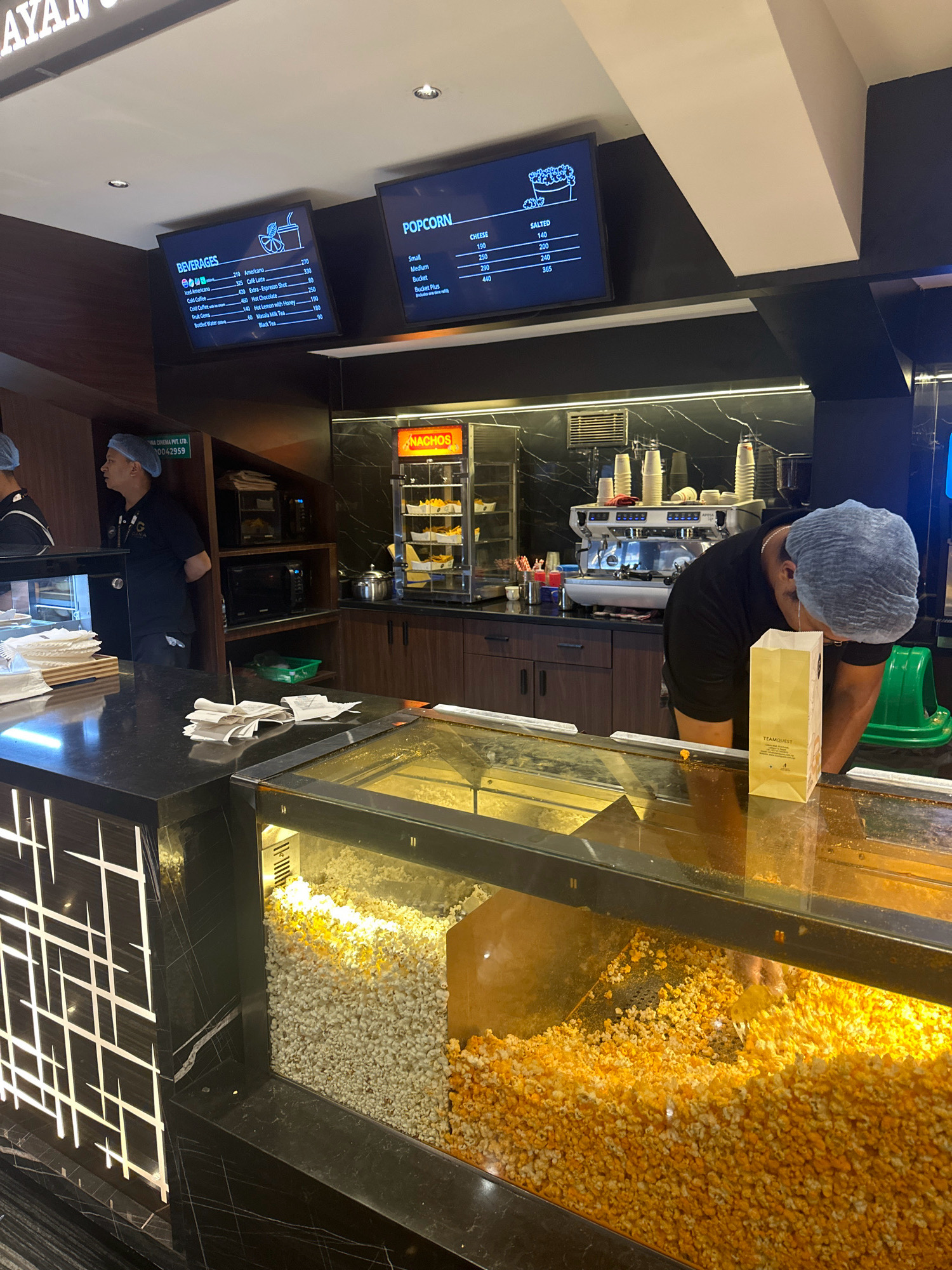 The image captures a concession stand inside a cinema in Lalitpur, Nepal. A male employee wearing a black t-shirt, black pants, and a blue hairnet is visible behind the counter, engaged in scooping popcorn into a paper bag. Another employee, also wearing a hairnet, is partially visible on the left side of the frame.
The counter features a glass display case filled with popcorn, with one section containing plain popcorn and the other with cheese-flavored popcorn. Several receipts are scattered on the counter. Above the counter, digital menu boards display beverage and popcorn prices. The beverage menu lists items such as Americano, Cold Coffee, and Black Tea, while the popcorn menu offers salted and cheese options in different sizes.
Behind the employees, a coffee machine and a display cabinet filled with nachos are visible, indicating a variety of food and drink options available. The overall environment suggests a typical cinema concession area, with clean surfaces and organized displays. The lighting is bright, illuminating the popcorn and the menu boards.