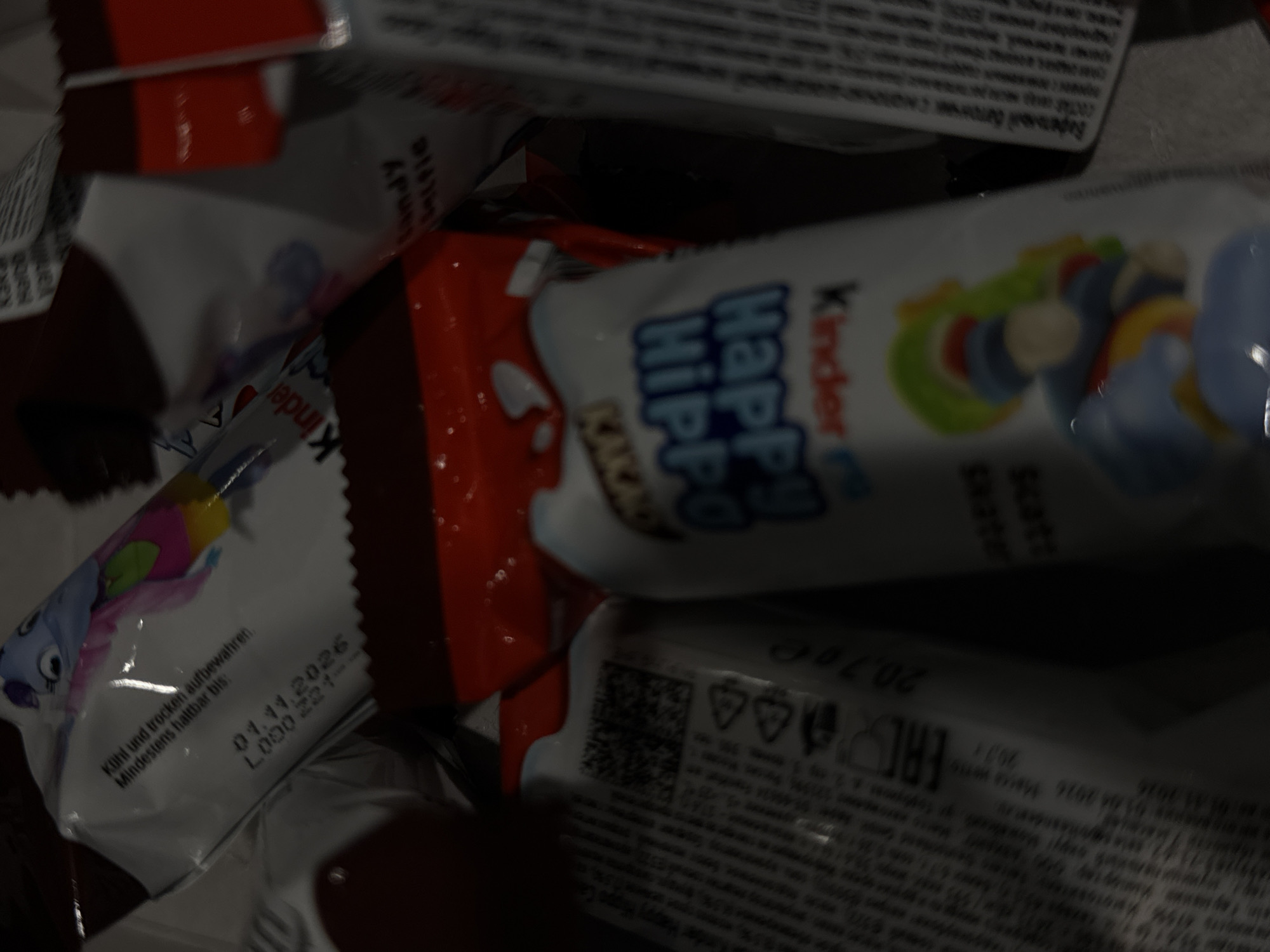 The image shows a close-up of several Kinder Happy Hippo snacks, some of which are in their original packaging. The packaging is white with colorful illustrations of cartoon characters and text in German and Russian. One package clearly displays the expiration date "01.11.2026". Another package has the text "Kinder Happy Hippo" in blue lettering. The lighting is dim, and the image is slightly blurred, suggesting it was taken in a low-light environment. There are no people visible in the image. The overall impression is of a pile of candy products. It is not possible to determine the location in Moscow, Russia, from this image alone.