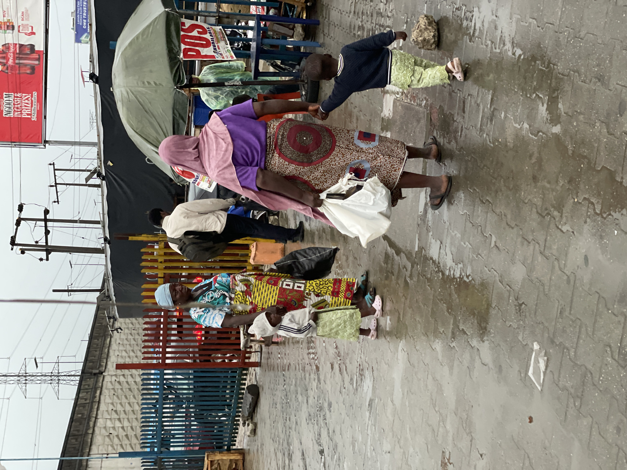 The image depicts a street scene in Lagos, Nigeria, likely after a rain. The ground is wet, reflecting light. Several people are visible, including two women and two children. One woman wears a purple top and a patterned skirt with a pink head covering, holding a child's hand. Another woman wears a colorful patterned top and a blue headwrap. A man walks in the background with a backpack. The scene includes street infrastructure like power lines, a red advertisement billboard promoting cash prizes, and a vendor stand with a green umbrella and "POS" (Point of Sale) sign. A fence separates pedestrians from the street.