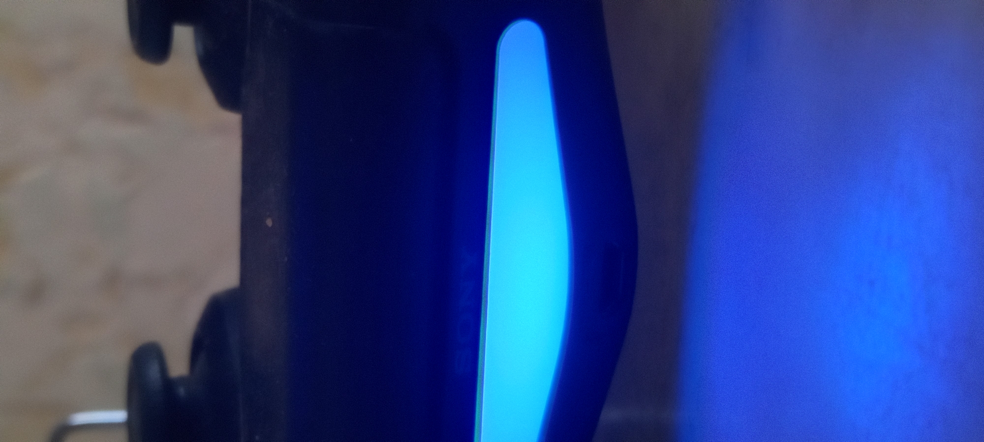 The scene depicts a close-up, front-facing view of a dark-colored Sony PlayStation DualShock 4 gaming controller, located in Kryvyi Rih, Ukraine. The controller's integrated light bar is prominently illuminated with a bright cyan-blue light, casting a darker blue reflection onto the surface immediately below it. The upper portion of the controller, including faintly visible "SONY" branding, is in soft focus, with two blurred, dark, rounded objects (likely analog sticks) partially visible in the upper-left and upper-right corners. A slender, bright, vertical object, possibly a metal hook or stand component, is discernible in the upper-left background. The background environment is heavily out of focus, showing a light-colored, textured surface. No individuals or active interactions are present within the frame.