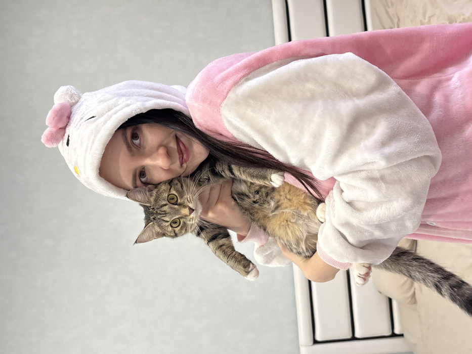 An adult female is depicted holding a domestic tabby cat in an indoor residential setting in Kazan, Russia. The female, exhibiting dark brown hair and reddish lipstick, wears a pink and white plush kigurumi featuring a white hooded design with cat ears, a pink bow, black embroidered eyes, and a yellow nose, indicative of a Hello Kitty motif. She supports the cat with both arms while looking directly at the camera with a subtle smile. The tabby cat, characterized by brown and grey striped fur, white paws, and yellow-green eyes, is also oriented towards the camera. The background includes a light blue-grey textured wall and a portion of a white, paneled bed headboard with light-colored bedding visible beneath.