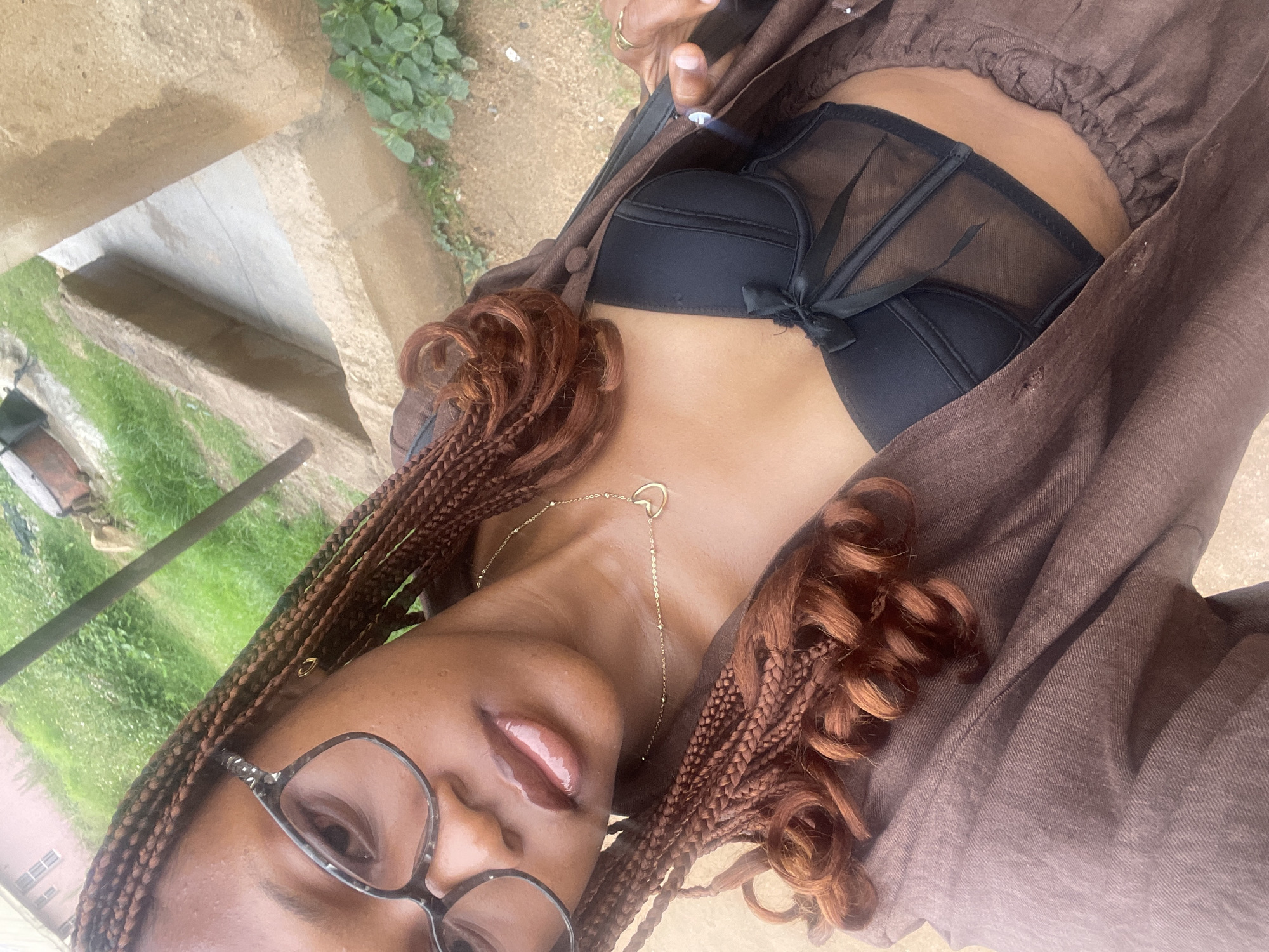 A young woman with braided hair and glasses is taking a selfie outdoors. She is wearing a brown open shirt over a black corset top with a bow. Her lips are glossy, and she wears a delicate gold necklace with a heart pendant. The background shows an outdoor environment with green grass and concrete structures, possibly in the city of Rafin Par, Nigeria.