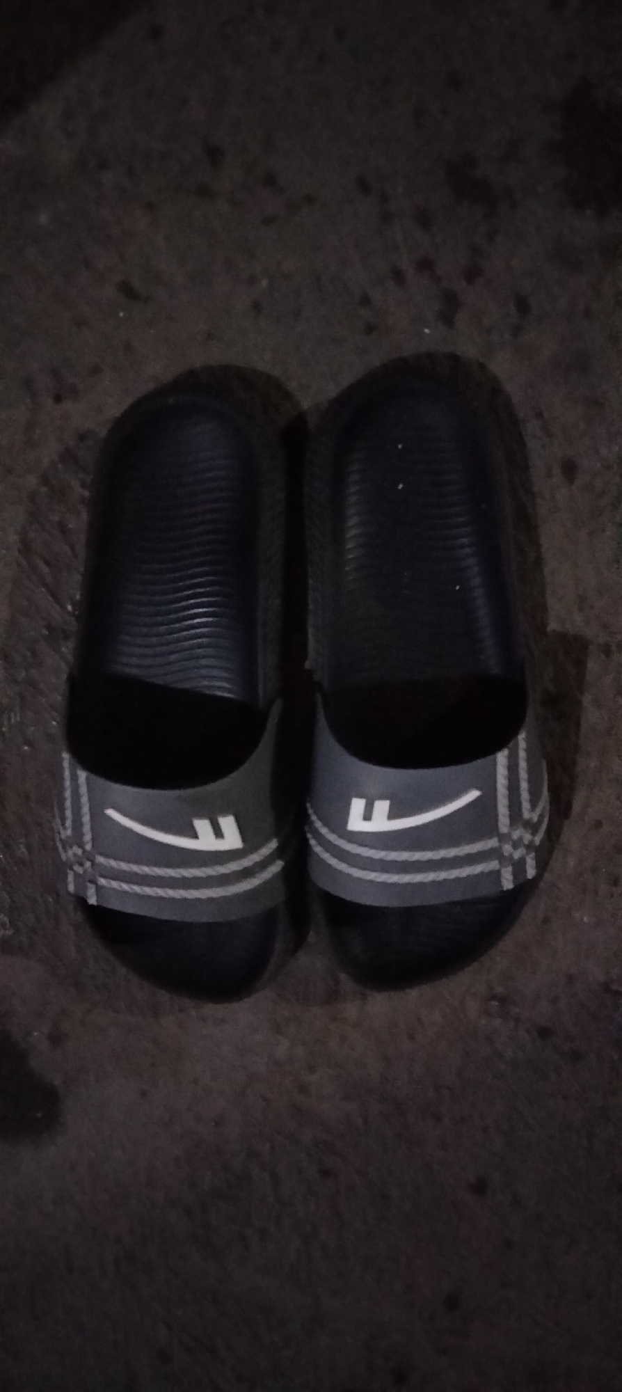 This is a top-down, full-frame shot of a pair of black slides with gray straps, resting on a dark, uneven surface, possibly asphalt or concrete, in the city of Serut, Indonesia. The slides are positioned side-by-side, with the left slide slightly ahead of the right. Each slide has a dark gray strap with two horizontal white stripes and a white graphic resembling a stylized "F" or a checkmark. The soles of the slides are black and appear to have a textured pattern. The surface they are on is dark and mottled, with some lighter specks and shadows cast by the slides, suggesting artificial lighting or the presence of overhead objects. The overall scene is dimly lit, with limited detail visible in the background. There are no people or any discernible action depicted.