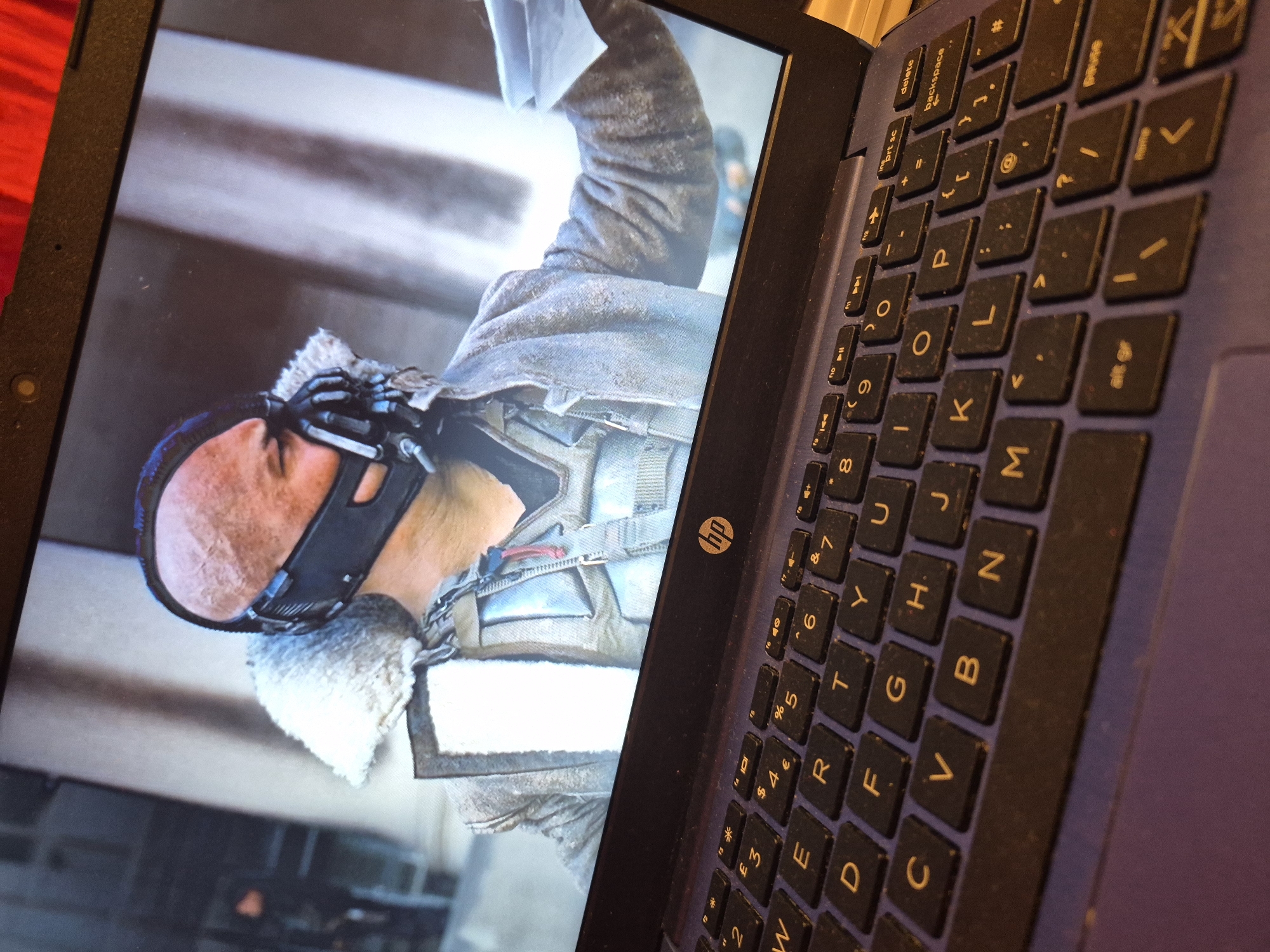 The image shows a close-up, angled view of a laptop screen displaying a scene from a movie, with a laptop keyboard in the foreground. The screen shows a close-up of a character wearing a distinctive black mask and a fur-lined jacket. The character is bald and appears to be male. The background on the screen is blurred, showing what appears to be architectural structures. The laptop keyboard is dark, with some keys visible, including letters like 'W', 'E', 'R', 'T', 'Y', 'U', 'I', 'O', 'P', 'A', 'S', 'D', 'F', 'G', 'H', 'J', 'K', 'L', 'Z', 'X', 'C', 'V', 'B', 'N', 'M', and various function keys. The overall lighting suggests an indoor environment with the laptop screen being the primary light source. There is no indication of the city of Sandwell, United Kingdom, in the image.