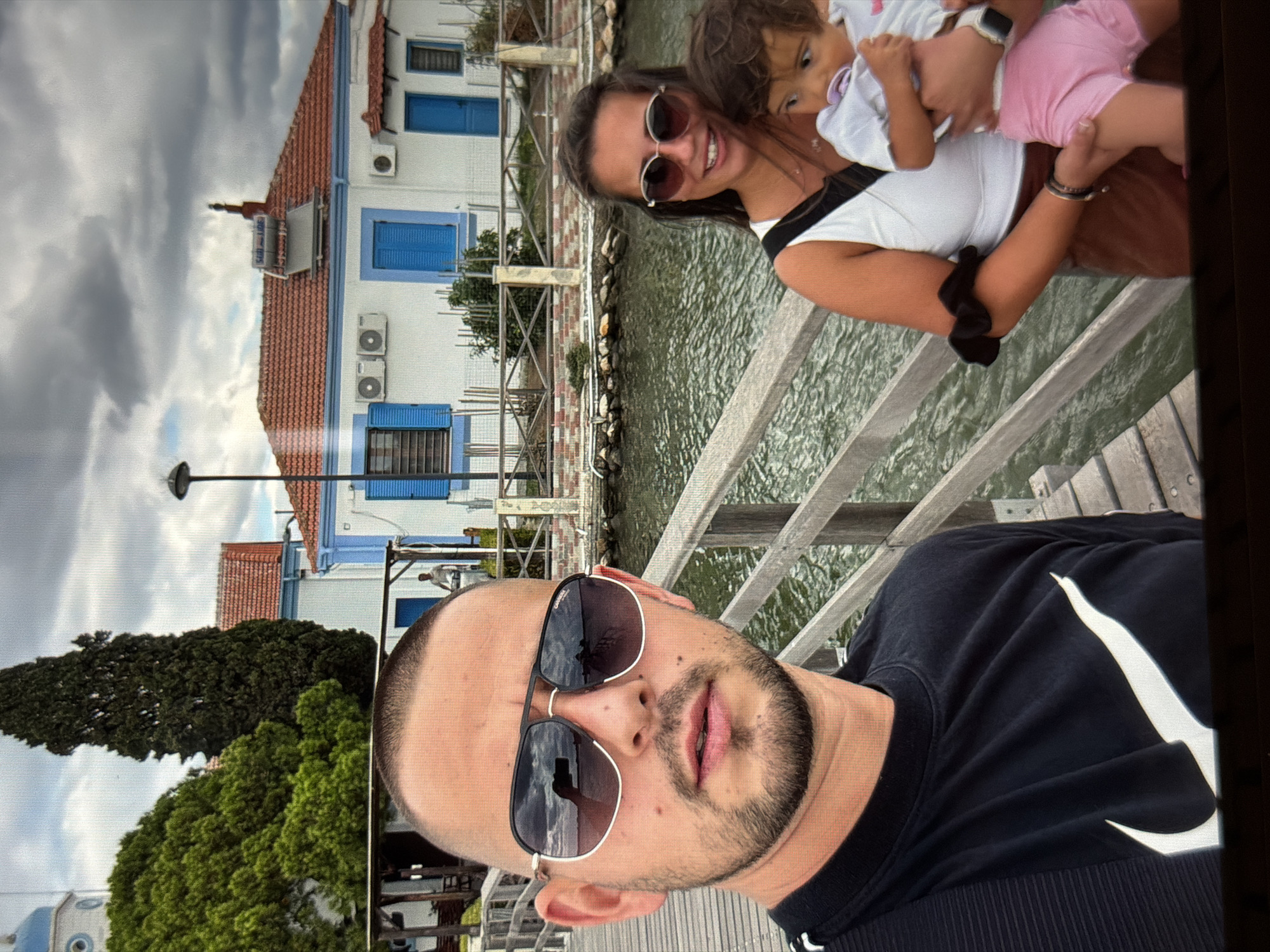 A family selfie captures a man, a woman, and a small child outdoors on what appears to be a wooden pier or walkway next to water. The man, positioned in the foreground, has a shaved head with short dark stubble, a beard, and a mustache. He wears dark sunglasses that reflect the cloudy sky and is dressed in a dark t-shirt featuring a prominent white Nike swoosh logo on the chest.
Behind him and to the right, a woman with long dark hair smiles broadly at the camera, wearing dark sunglasses and a white top with black detailing. She holds a young child, likely a toddler, who has curly light brown hair, a pacifier in their mouth, and is dressed in a light-colored top and pink shorts. The child looks off to the side, away from the camera.
The background features calm water, possibly a canal or bay, bordered by a decorative wall. Across the water, several two-story buildings with white facades, blue window shutters, and terracotta tile roofs are visible. Air conditioning units are mounted on their exterior walls, and a solar water heater marked "500 L" can be seen on one rooftop. Lush green trees and foliage are interspersed among the buildings. A black streetlight pole stands to the left, and a partial view of a blue-domed structure with white details is visible in the far left background.
The setting suggests a leisurely daytime outing under an overcast sky. The architecture, including the blue shutters, terracotta roofs, and the visible blue dome, aligns with coastal regions, consistent with the location context of Burgas, Bulgaria. The woman is smiling, conveying a pleasant atmosphere.