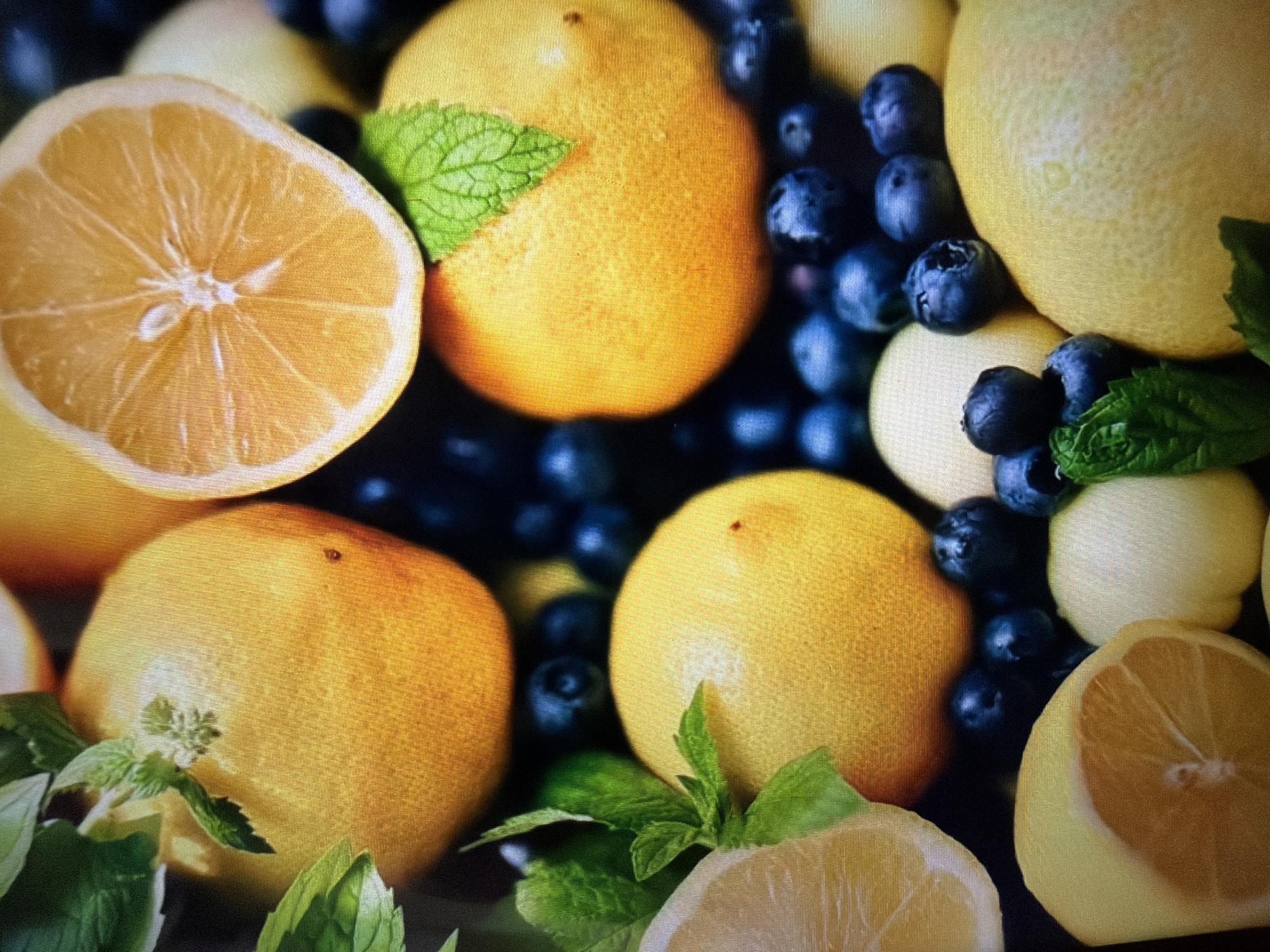 A close-up, overhead shot showcases a vibrant arrangement of fresh fruits and mint leaves. Several whole lemons, some sliced in half, are interspersed with dark blue blueberries. A few sprigs of green mint add a fresh contrast to the bright yellow of the lemons and the deep blue of the berries. The fruits are artfully placed, creating a visually appealing still life composition. The background is dark and blurred, drawing attention to the foreground elements. There are no people visible in the image, and no indication of a specific location in Moscow, Russia. The image appears to be a photograph of produce, possibly for a culinary or decorative purpose.