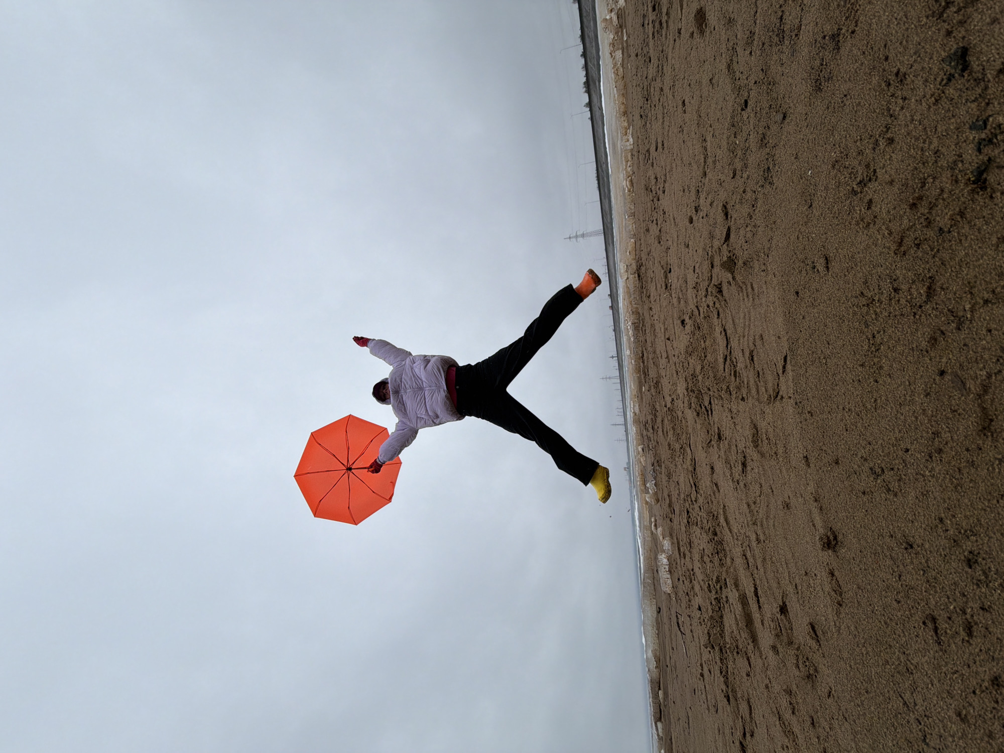 An image, rotated ninety degrees clockwise from its intended orientation, captures a person mid-air against an overcast sky and sandy terrain. The main subject is an individual, positioned horizontally as if suspended or flying, with arms and legs outstretched. They wear a light-colored (possibly white or light purple) puffy jacket, dark trousers, a bright orange glove on one hand, and distinctive yellow footwear on the other. In their left hand, they hold an open, vibrant orange umbrella.
The background is dominated by a vast, plain grey sky, indicative of an overcast day. Below, the ground is a rough, sandy or dirt-like expanse, possibly a beach or a muddy shoreline, extending towards a distant horizon. A faint, light-colored strip, potentially ice, snow, or shallow water, marks the edge of the land. Along this distant line, several faint vertical structures, resembling utility poles or power lines, are visible against the muted sky.
The activity depicted is a dynamic, almost acrobatic pose, suggesting a staged or playful moment where the person appears to defy gravity, perhaps mimicking flight or being carried by the wind. The vibrant umbrella and footwear stand out against the otherwise muted color palette of the sky and ground. There is no visible text in the image. The setting appears to be an outdoor, expansive landscape, consistent with the provided location of Chaikovsky, Russia.