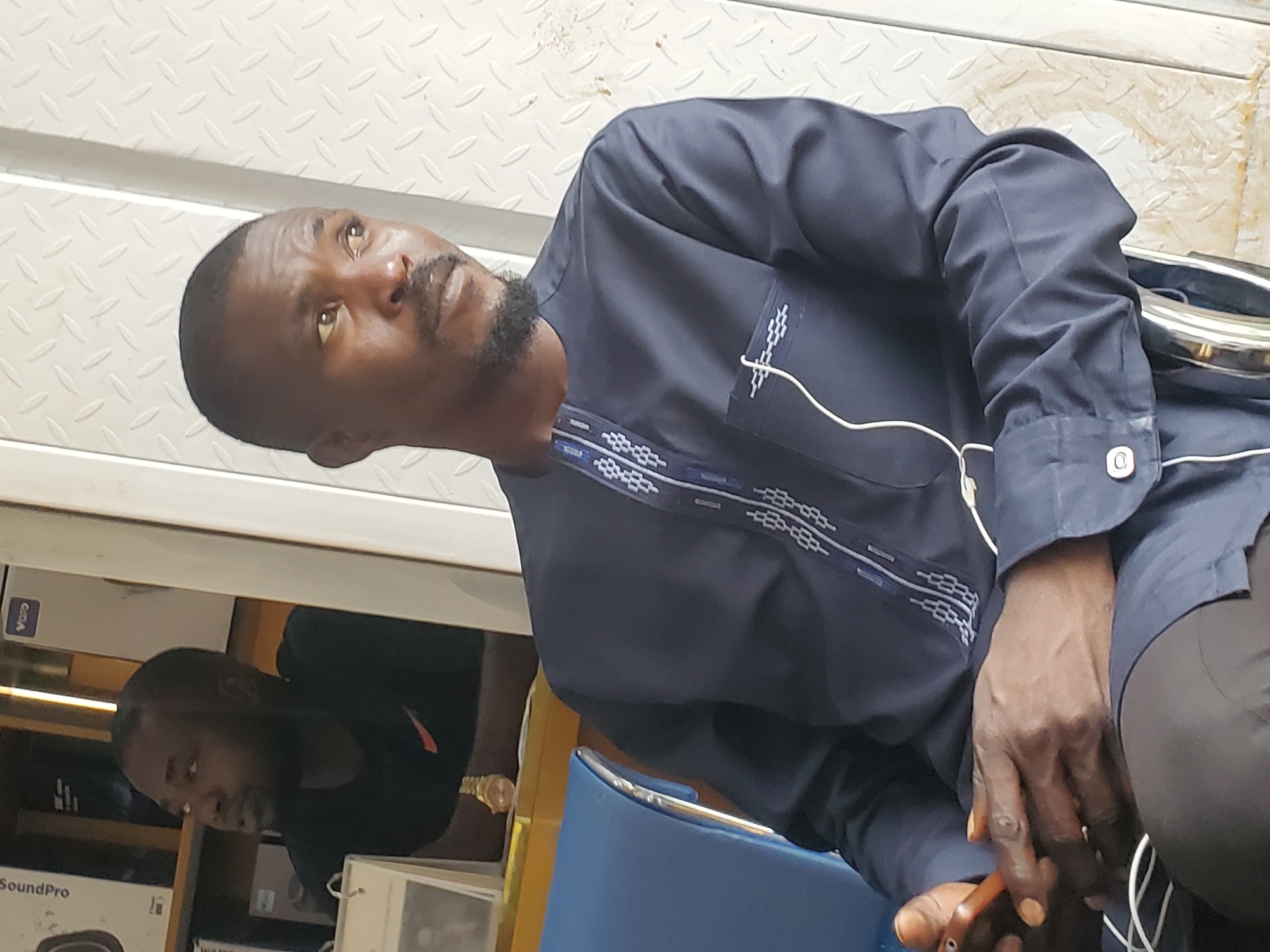 The image shows a man sitting on a blue chair, looking to his right. He is wearing a dark blue traditional Nigerian outfit with intricate white embroidery along the neckline and on the left breast pocket. The man has a goatee and is holding a mobile phone in his hands. In the background, a reflection shows another man in a black t-shirt looking to his left, with shelves of electronics behind him. The environment appears to be an indoor space with a corrugated metal wall behind the man. This scene could be interpreted as being in a shop or a waiting area, potentially in Jos, Nigeria, given the traditional attire and common electronics found in such shops in West African cities.