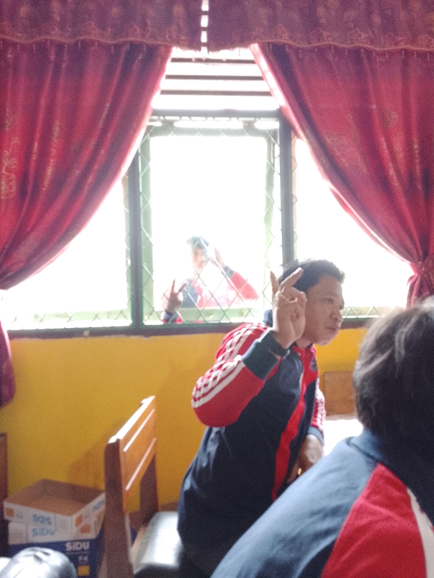 The image depicts an indoor scene, likely a classroom or meeting room, with yellow walls and large red curtains framing a window. The window offers a view of another person outside, visible through a green lattice. In the foreground, a young man wearing a navy blue and red tracksuit is seated and making a "peace" or "victory" gesture with his right hand. Another person, mostly out of frame, is seen from behind, also wearing a similar tracksuit with red and blue accents. A wooden chair and a cardboard box labeled "SIDU" are visible in the lower left portion of the frame. The lighting suggests it is daytime, with bright light filtering through the window. The overall atmosphere appears casual and possibly informal, given the clothing and gestures. There is no discernible indication of the specific location in Jepara, Indonesia.