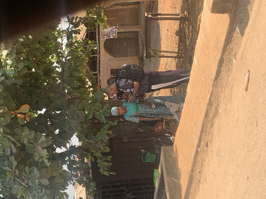 The scene depicts a group of four individuals standing under a large deciduous tree on an unpaved, reddish-brown dirt ground in Egbedi, Nigeria, during daylight hours.
In the center-left, a woman in a teal-green patterned long dress and matching headwrap holds a small, dark-skinned child in her left arm. She is smiling and looking towards a man to her right. The child, wearing dark clothing and light sandals, is also looking towards a mobile phone.
To the right of the woman and child, a man with a shaved head wears a black and white striped short-sleeved shirt, dark pants with a white side stripe, and a black backpack. He is holding a black mobile phone, presenting its screen to the woman and child. Partially obscured behind this man is another man in a dark shirt.
The group appears to be engaged in viewing content on the mobile phone.
In the left background, a dark, corrugated structure with a barred window is visible. A green plastic bucket and two green crates are positioned near this structure. In the right background, a light-brown, partially dilapidated brick building with two arched openings and exposed wooden roof beams stands. A wooden pole with a poster is affixed in front of this building.
A concrete curb or low wall runs across the midground from left to center. A simple, four-legged wooden table is situated in the right midground. A small, light-colored piece of debris lies on the dirt ground in the foreground.