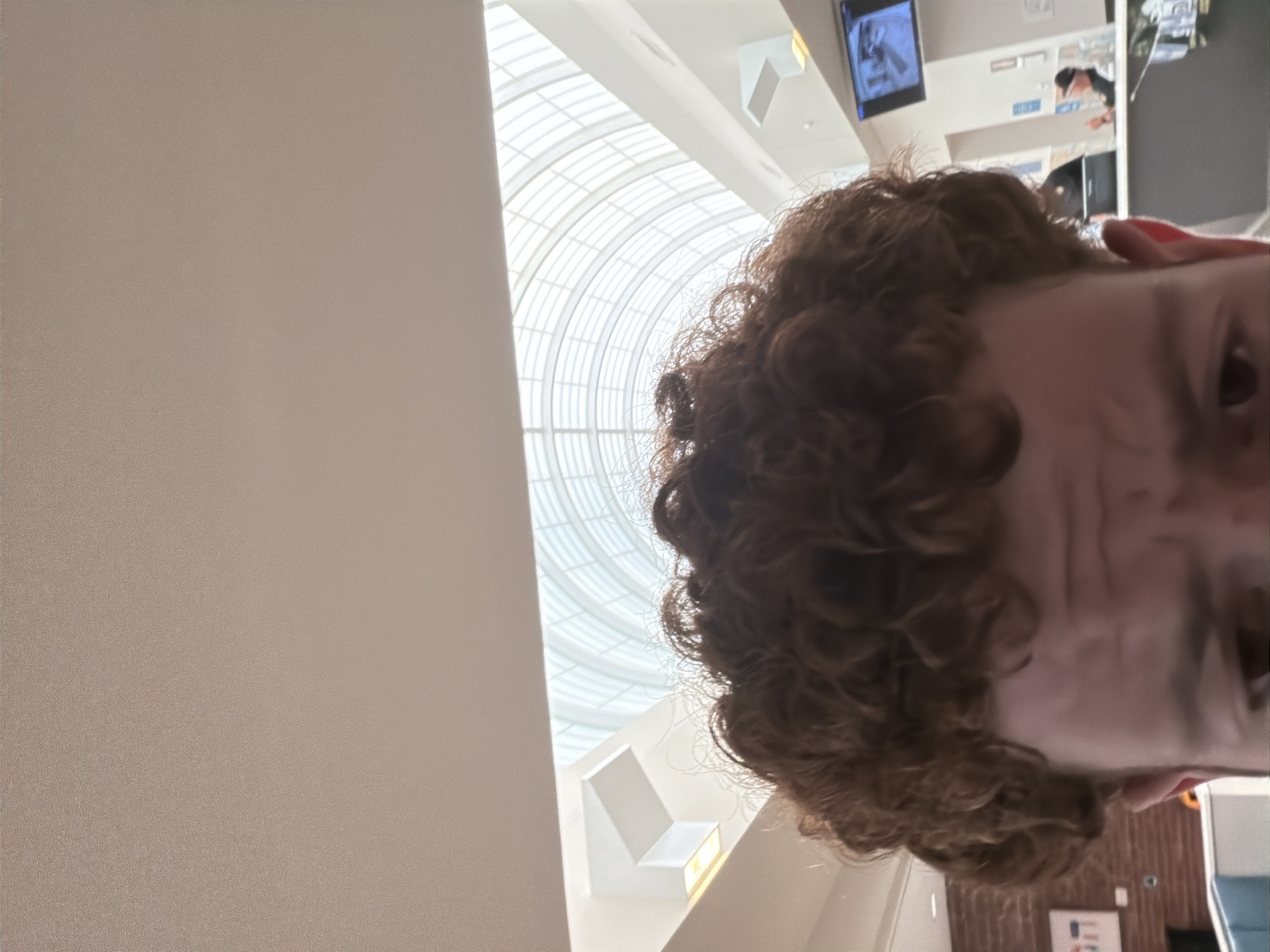 This is a close-up, low-angle photograph of the top of a person's head and face, with their curly, reddish-brown hair filling the foreground. The individual appears to be looking directly at the camera with a slightly furrowed brow. In the background, a modern, brightly lit interior space is visible, featuring a large, arched glass ceiling that lets in natural light. The architecture suggests an indoor mall or a similar public atrium.
Further back, a reception desk area can be seen, with at least two individuals present. One person is wearing a dark uniform, possibly security, and another individual is wearing a hat. A security camera is mounted on the wall above the desk, and a monitor displays an image, likely from a surveillance feed. To the left of the reception area, a brick wall and a framed piece of art or signage with Hebrew lettering are visible. The overall environment is clean and contemporary.
The photograph was taken at the Beachwood Place mall in Beachwood, Ohio, United States of America.