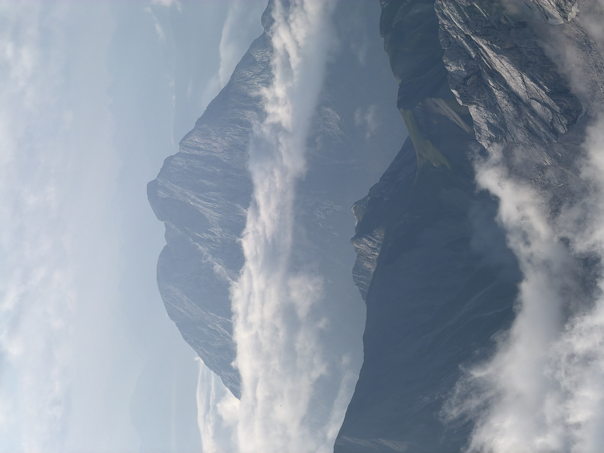 This is a vertical, medium shot photograph of a mountain range in Grainau, Germany. The scene is dominated by layers of mountains and clouds. The uppermost, most distant mountain is a muted grey with a textured surface, appearing somewhat hazy. Below it, a larger, more rugged mountain peak emerges from a band of white clouds, its slopes appearing steep and rocky. Further down, another mountain range is visible, its dark blue-grey slopes marked by sharp ridges and lighter colored scree or rock formations. The foreground features another mountain with a textured, grey, rocky surface, partially obscured by wispy white clouds. Green vegetation is visible on some of the lower mountain slopes. The sky above is a hazy blue with scattered white clouds. The overall impression is one of a vast, rugged mountain landscape partially veiled by atmospheric conditions.