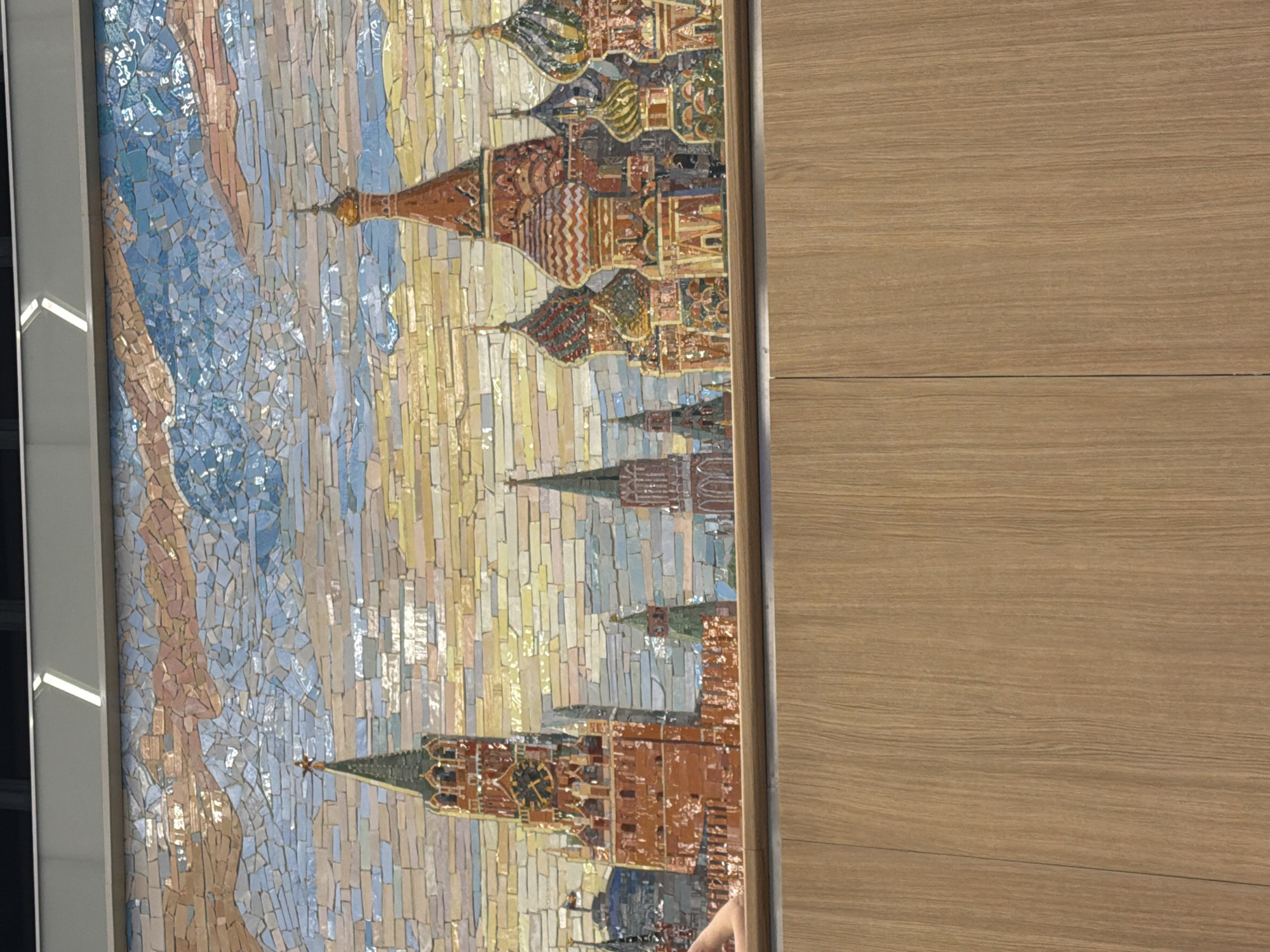 The image shows a mosaic depicting a cityscape, likely Red Square in Moscow, Russia. The mosaic features detailed renderings of iconic Russian architectural elements, including a prominent clock tower resembling the Spasskaya Tower and several onion-domed churches characteristic of St. Basil's Cathedral. The sky above is rendered with a mosaic of various shades of blue, brown, and orange, suggesting a sunrise or sunset. In the lower left corner of the mosaic, a portion of a person's hand and arm is visible, suggesting someone is observing or interacting with the artwork. The mosaic is mounted above a wooden paneling, which occupies the lower portion of the image.