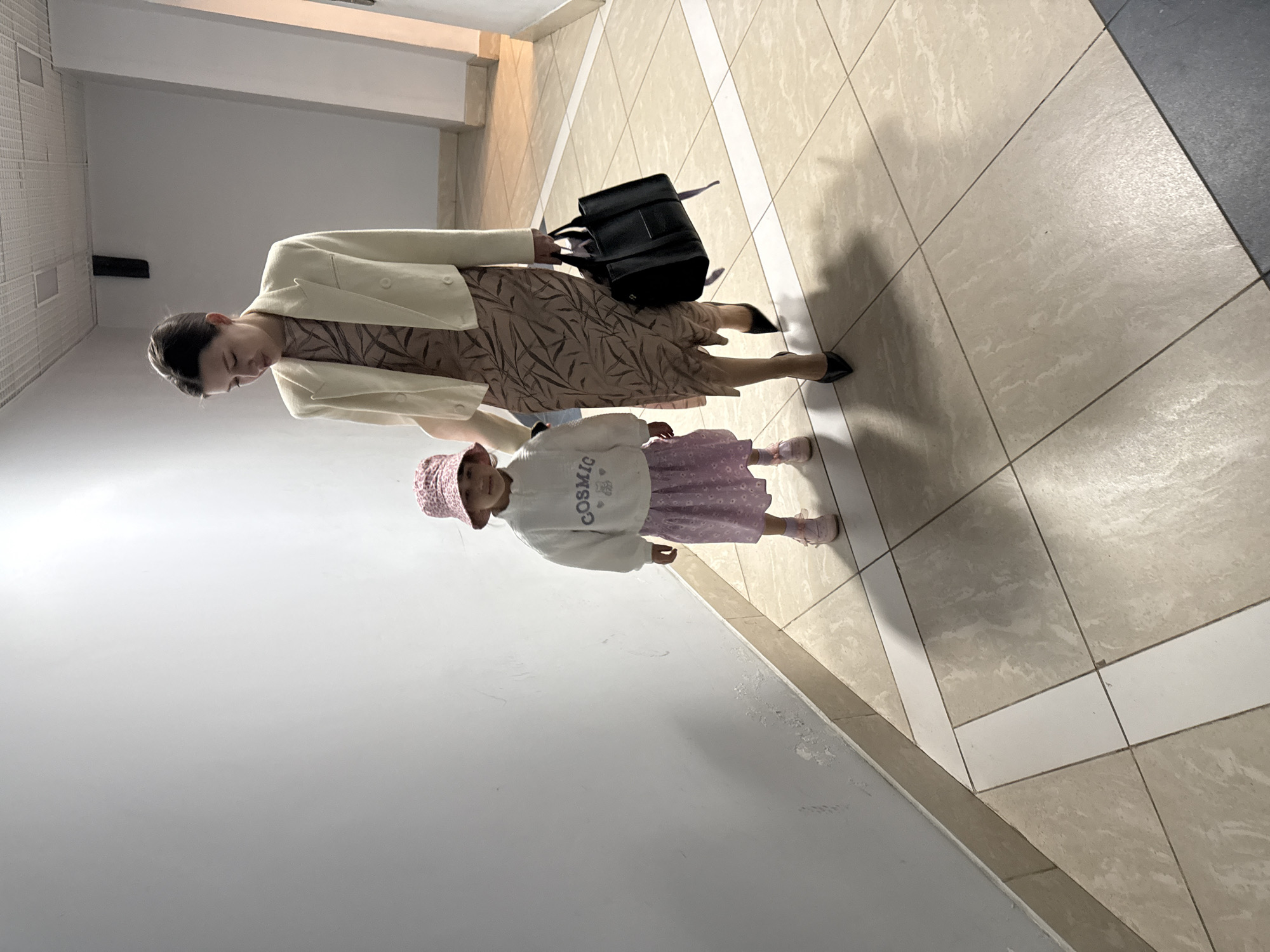 The image, rotated 90 degrees counter-clockwise, captures a woman and a young child in an indoor corridor or hallway, likely within a public or commercial building.
The adult, positioned on the right side of the frame, has dark hair styled neatly. She wears a light-colored (off-white or cream) blazer over a long-sleeved, knee-length dress with a subtle tan and brown leaf-like pattern. She carries a black handbag in her right hand and is wearing dark, pointed-toe heels. Her head is tilted downwards, and her gaze is directed away from the viewer.
To her left, a young child, likely a girl, stands facing the camera with a neutral expression. She is dressed in a light-colored (white or cream) sweatshirt, a pink patterned skirt, and a pink bucket hat. The word "COSMIC" is visible in dark letters on the front of her sweatshirt. She wears light pink shoes.
The environment features light-colored, square floor tiles (beige/off-white) with dark gray grout lines. White strips are incorporated into the tile pattern, creating geometric divisions on the floor. A section of darker gray or black flooring is visible on the far right. The walls are light-colored, with a textured, paneled section on the left. Artificial lighting is present, casting shadows of the subjects on the floor, suggesting an indoor setting at an indeterminate time of day. The scene depicts them standing or moving through this space.