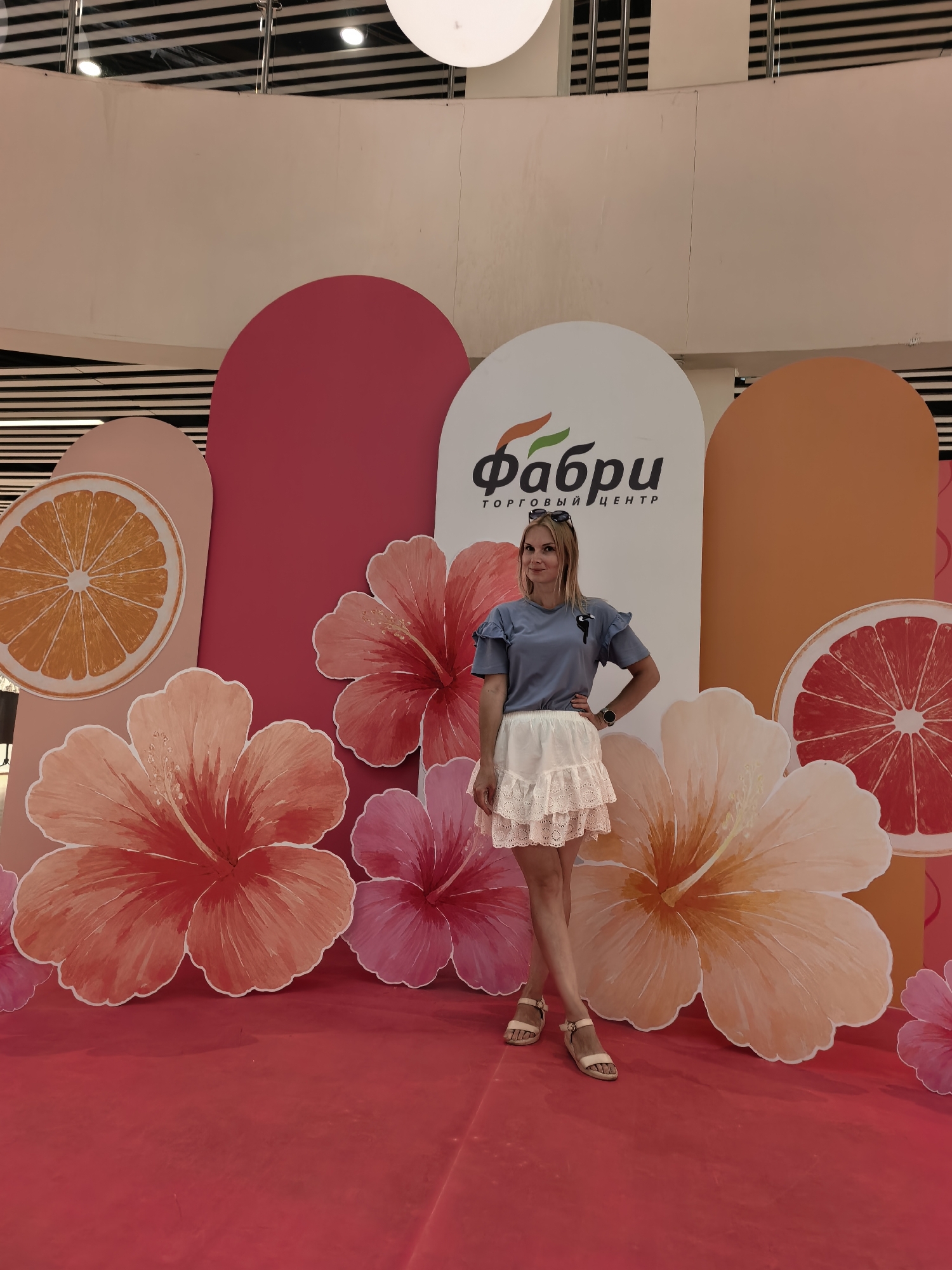 A blonde woman stands in front of a colorful backdrop featuring oversized tropical flowers and fruit slices, with a logo that reads "Фабри" in Russian. The woman is wearing a light blue t-shirt with ruffled sleeves and a white lace skirt, paired with beige sandals. She has her left hand on her hip and is looking at the camera. The backdrop appears to be a promotional display, likely in a public space such as a shopping center. The "Фабри" logo suggests this location is "Fabri Shopping Center". Given the context provided in the prompt, the image was taken in Sterlitamak, Russia.
