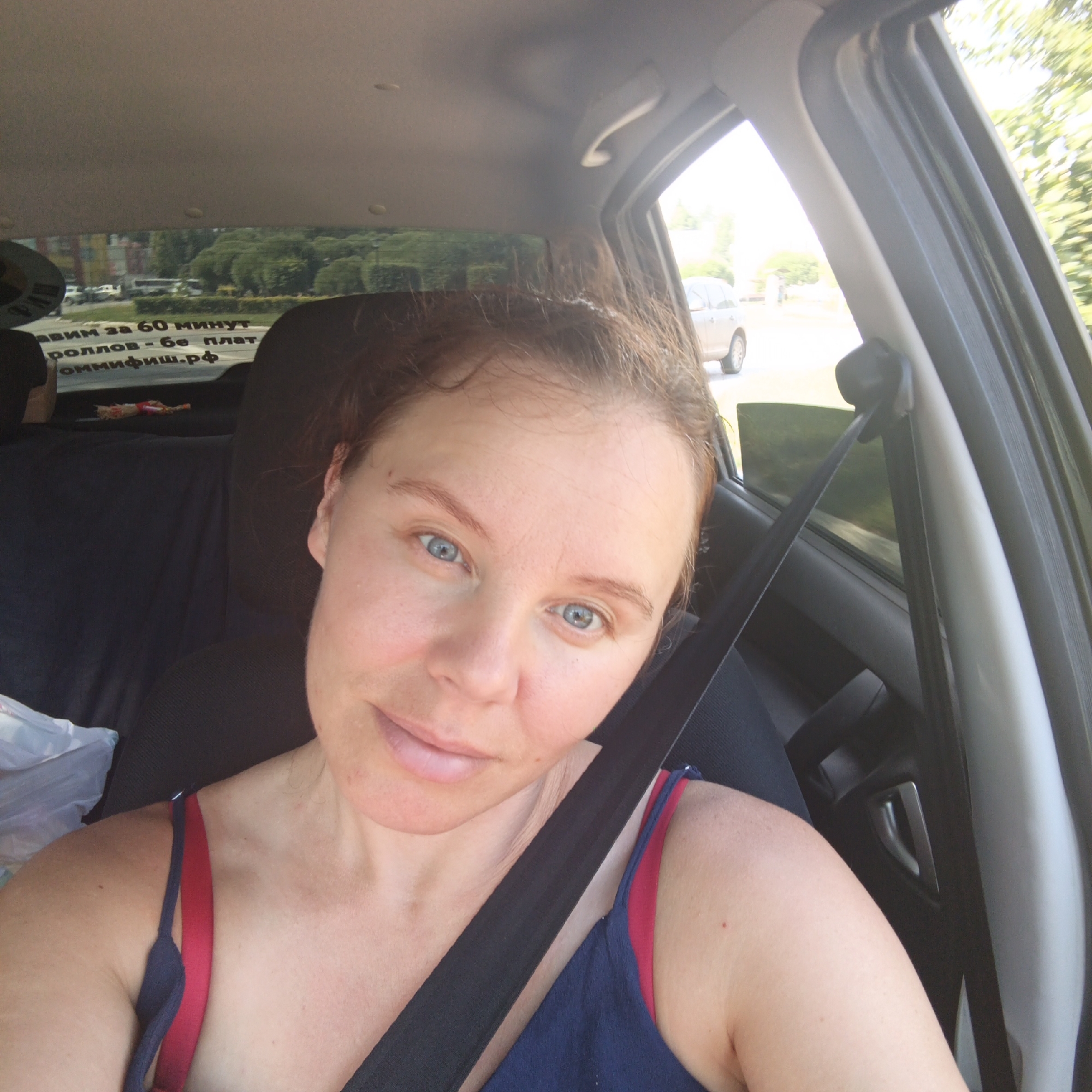 The image shows a woman with light skin and blue eyes taking a selfie inside a car. She has light brown hair. She is wearing a blue and red tank top. A black seatbelt is across her chest. In the background, through the car window, trees and buildings can be seen. Text is visible on the rear window of the car, written in Russian. The text reads "ЗАВИМ ЗА 60 МИНУТ РОЛЛОВ - БЕ ПЛАТ ОМНИФИШ.РФ". This translates to "We deliver in 60 minutes Rolls - free Omnifish.rf". The context of Chaikovsky, Russia is not directly evident from the image itself but is provided as a location.