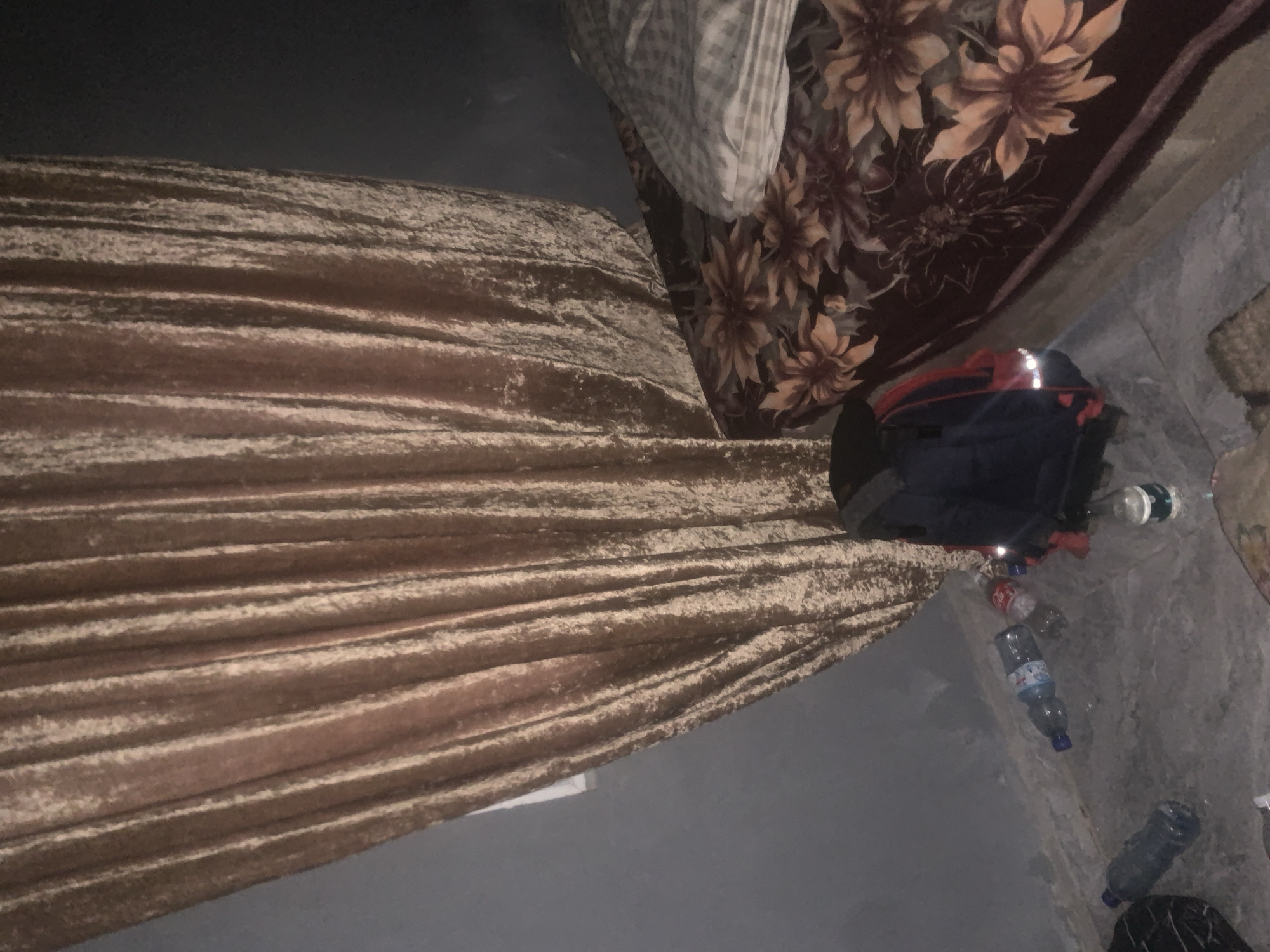 The image depicts an indoor scene with a bed covered in a dark brown floral-patterned blanket. A light-colored pillow with a checkered pattern rests on the bed. In the foreground, several plastic bottles are scattered on the floor, including one clear bottle with a blue cap, another clear bottle with a white label, and a red and white can. A dark blue backpack with red accents is positioned on the floor near the bottles. A textured, light brown curtain hangs prominently on the left side of the image, partially obscuring a gray wall. The overall environment appears to be a bedroom or living space. This scene is observed in Jalingo, Nigeria.