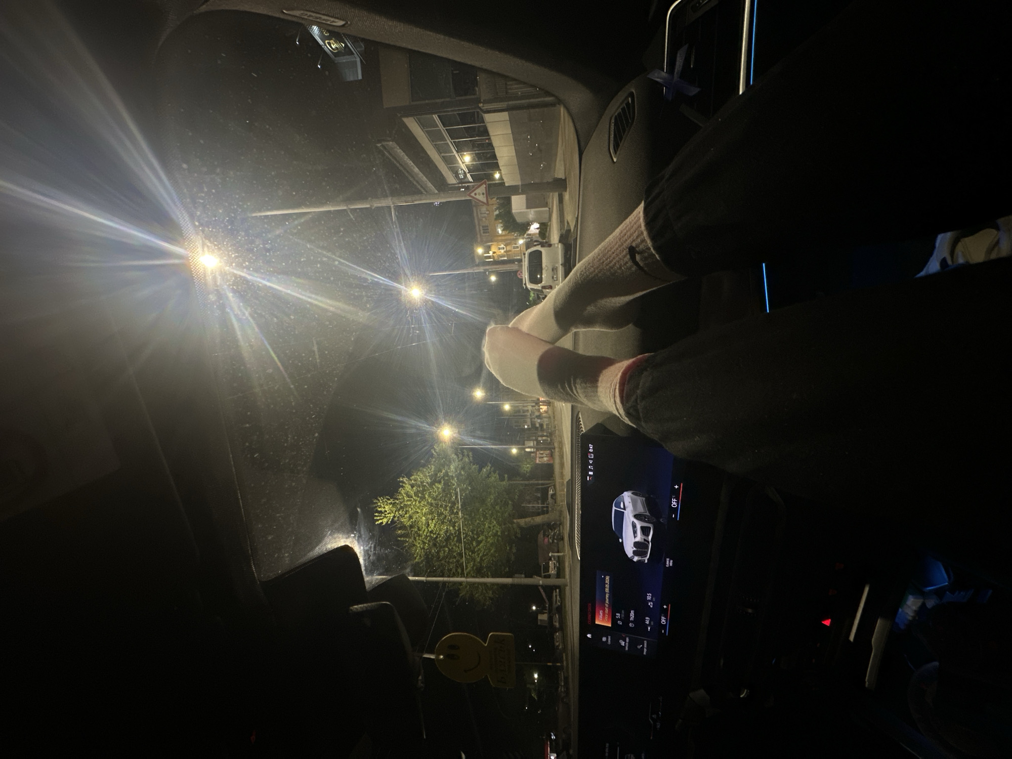 The image is taken from inside a car at night, providing a view of the road and the interior of the vehicle. The feet, clad in white socks, are placed on the dashboard. Through the windshield, a street is visible, illuminated by streetlights. The dashboard is visible, featuring a digital display showing information, including a date and time, the duration of a journey (1h20m), and other data, as well as what appears to be a graphic of a car. A small smiley face object is seen hanging from the rearview mirror.