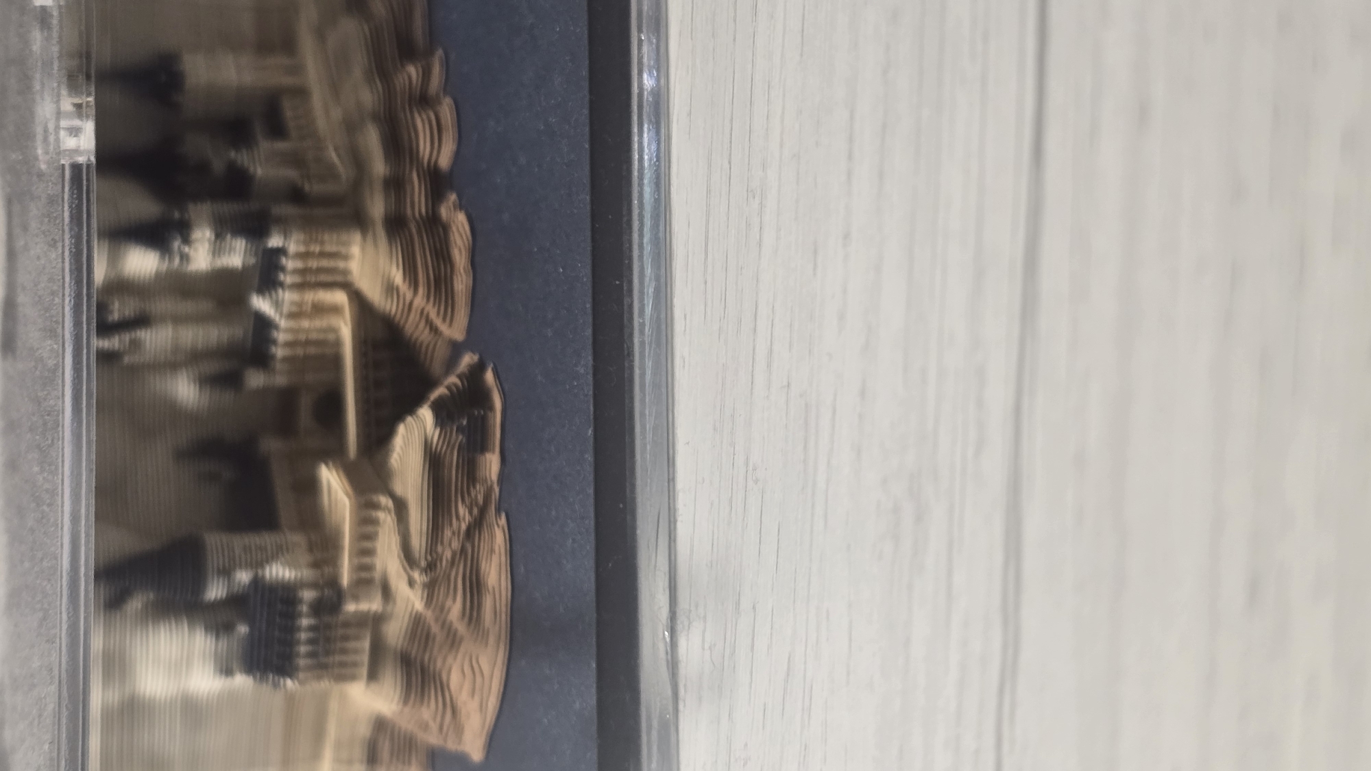 The image shows a layered model of a castle, possibly Hogwarts from Harry Potter, enclosed in a clear plastic case. The castle model is made of what appears to be cardboard or paper, with distinct layers creating a three-dimensional effect. The castle has multiple towers with pointed roofs and a detailed structure. The base of the model is dark blue, and the surrounding area of the image is out of focus, showing a light-colored, textured surface, likely a wooden table. There is no indication of the city of Ukhta, Russia in this image.