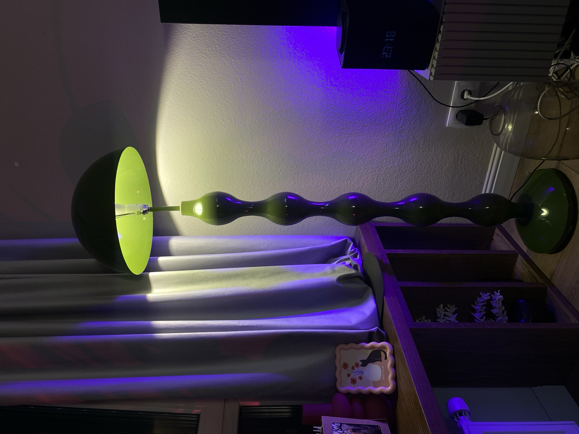 A green mushroom-shaped floor lamp with a bright yellow interior is positioned in the center of the frame, casting a yellowish light on the wall behind it. The lamp has a tall, segmented green base with a series of bulbous sections. To the left of the lamp, grey curtains are illuminated by purple and white light. On a wooden shelf beneath the curtains, there is a pink scalloped frame containing a picture of a black cat and flowers in a vase, and a red vase. To the right of the lamp, a black digital clock displays "23:18" and is plugged into a white electrical outlet next to a black plug. A ribbed metallic object is situated next to the clock, and a large glass carafe with a white cord lies on the floor in front of the outlet. The floor is made of wooden planks. The scene appears to be an indoor domestic setting, possibly a living room or study. The overall ambiance is characterized by colored lighting. This image does not depict the city of Moscow, Russia, or any identifiable landmarks associated with it.