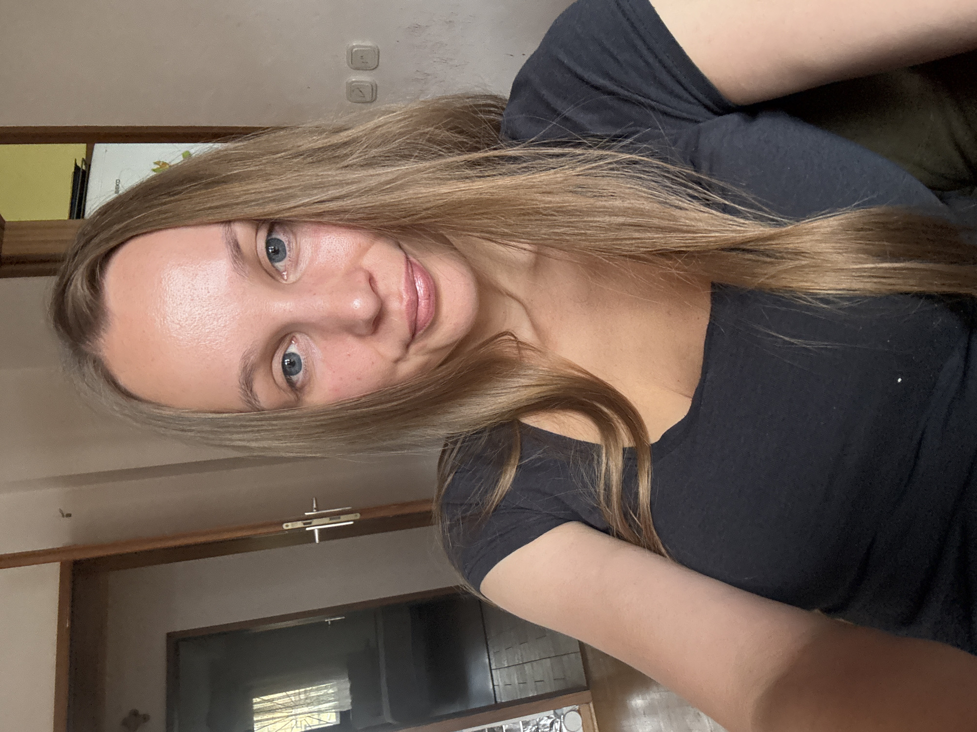 A young Caucasian woman with long, straight blonde hair and blue eyes smiles slightly at the camera. She is wearing a dark grey t-shirt. The background appears to be an indoor residential setting, possibly a home in Überherrn, Germany, with doorways, a wall, and what looks like a poster on the wall. The lighting suggests it is daytime.