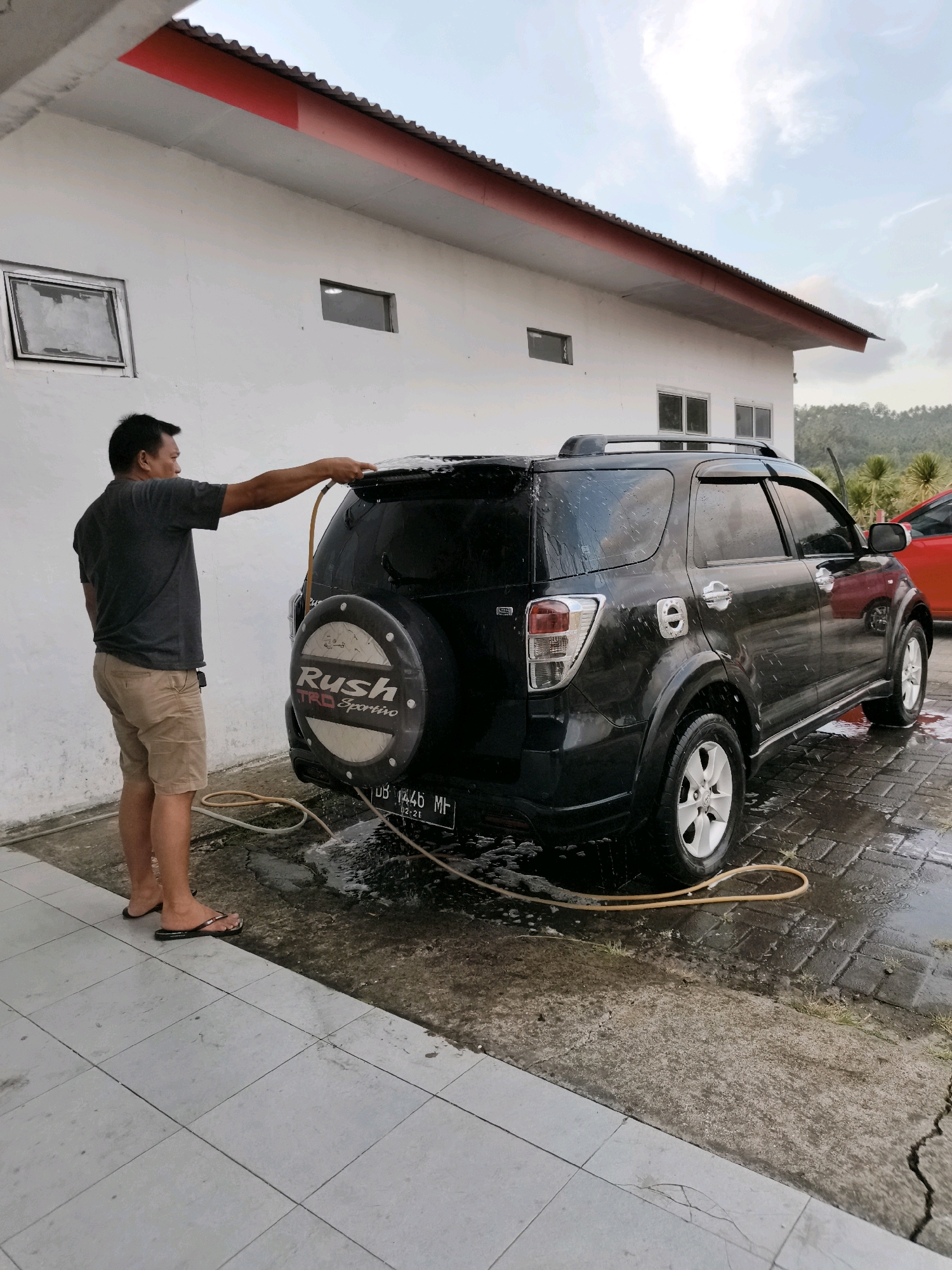 A middle-aged Asian man is washing a black Toyota Rush SUV in front of a white building with a red roof trim. The building has two windows and a covered overhang. The man is wearing a dark gray t-shirt and khaki shorts, and flip-flops. He is holding a yellow hose with a nozzle, spraying water on the rear of the SUV. The SUV is wet and soapy, with water running down its sides and onto the wet brick-paved ground. A spare tire cover with the "Rush TRD Sportivo" logo is visible on the back of the SUV, as well as a license plate that reads "DB 1446 MF". A red car is partially visible behind the black SUV. The scene is set in what appears to be a residential or semi-commercial area, possibly in Tombatu Dua, Indonesia, with some vegetation and trees visible in the background against a cloudy sky.