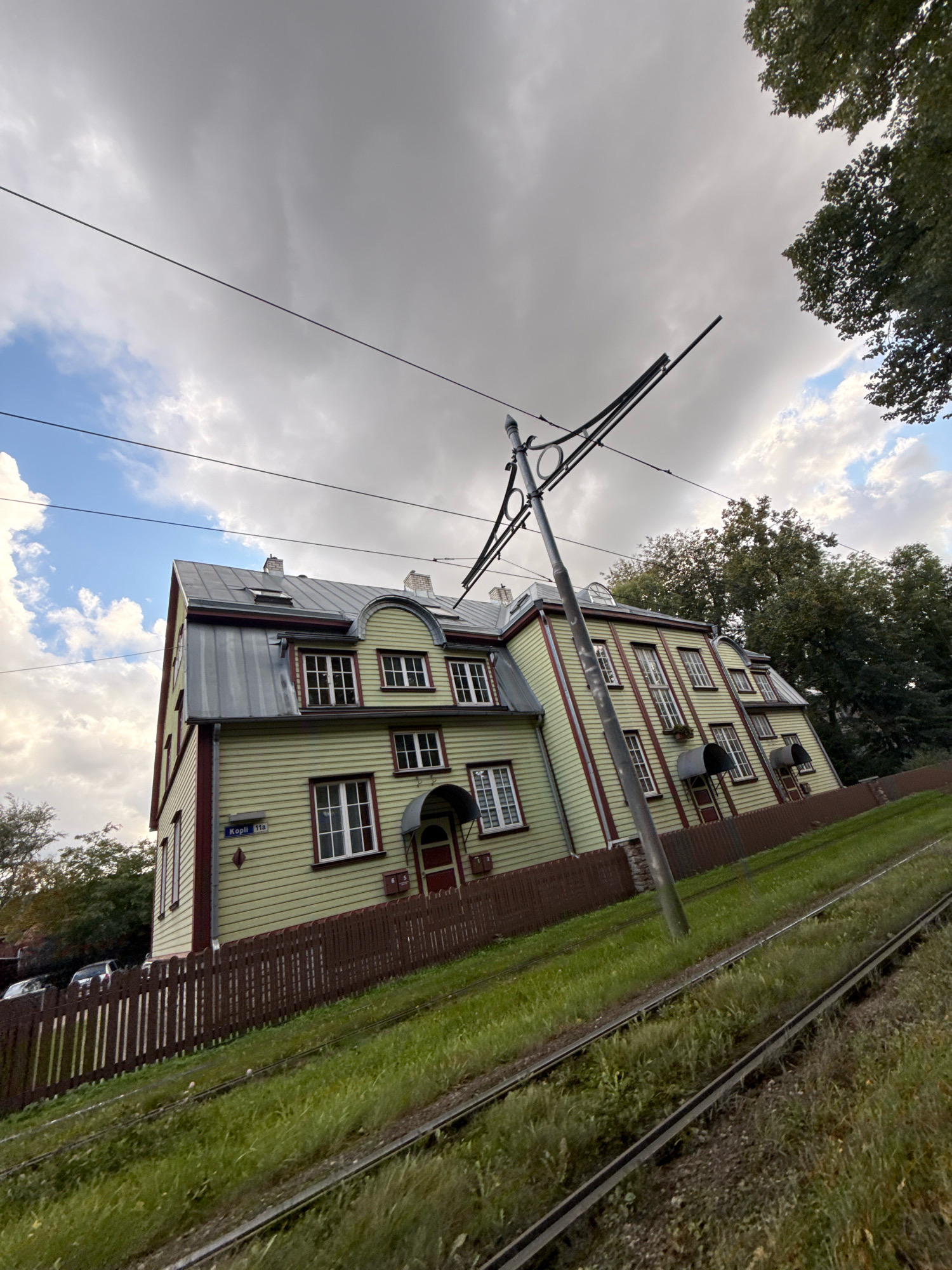 The image shows a long, light green wooden building with a dark red trim, featuring multiple dormer windows and chimneys. The building is situated on a slope, with tram tracks running parallel to it in the foreground. A dark brown wooden fence runs along the front of the property. There are power lines overhead, supported by a metal pole. The sky is cloudy with patches of blue. A street sign visible on the building reads "Kopli 11a," indicating the location is Kopli Street, number 11a. The overall scene depicts a residential or mixed-use building in a somewhat tilted perspective, likely in Tallinn, Estonia, given the street name.