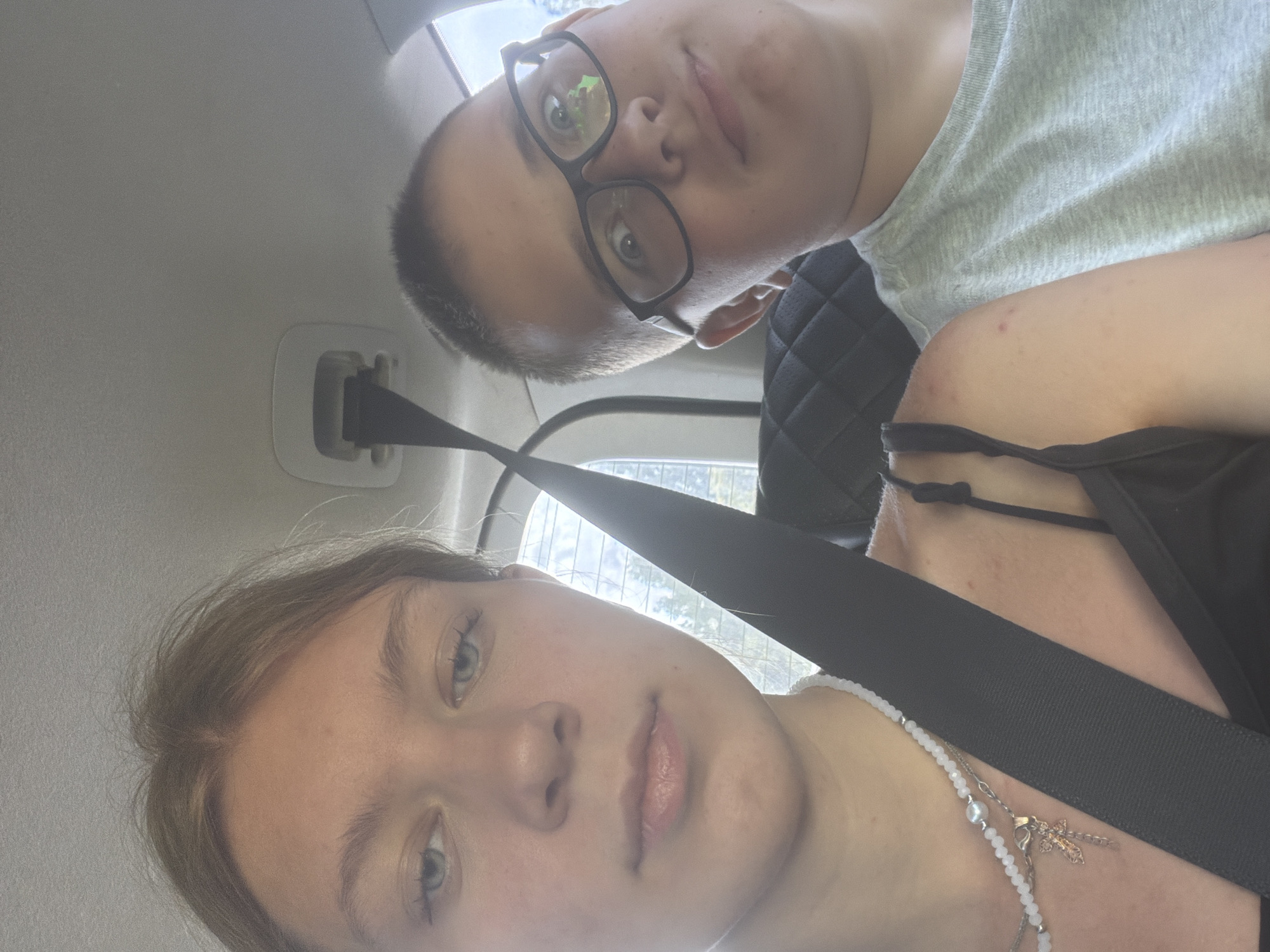 Two individuals, a female and a male, are visible in the foreground of a selfie taken inside a vehicle. The female, on the left, has light-colored hair pulled back and light blue eyes. She is wearing a white beaded necklace with a cross pendant and a black strap top. The male, on the right, has short dark hair and is wearing glasses and a light gray t-shirt. He appears to be looking at the camera with a surprised expression. Both individuals are wearing seatbelts, indicated by black straps across their chests. The background shows the interior of the car, including a quilted seat and a portion of the car's ceiling. Outside the car window, trees are visible, suggesting an outdoor setting. The scene depicts a candid moment within a vehicle, potentially during a journey. The location of Nadezhdino, Russia is not depicted in the image and is extraneous information.