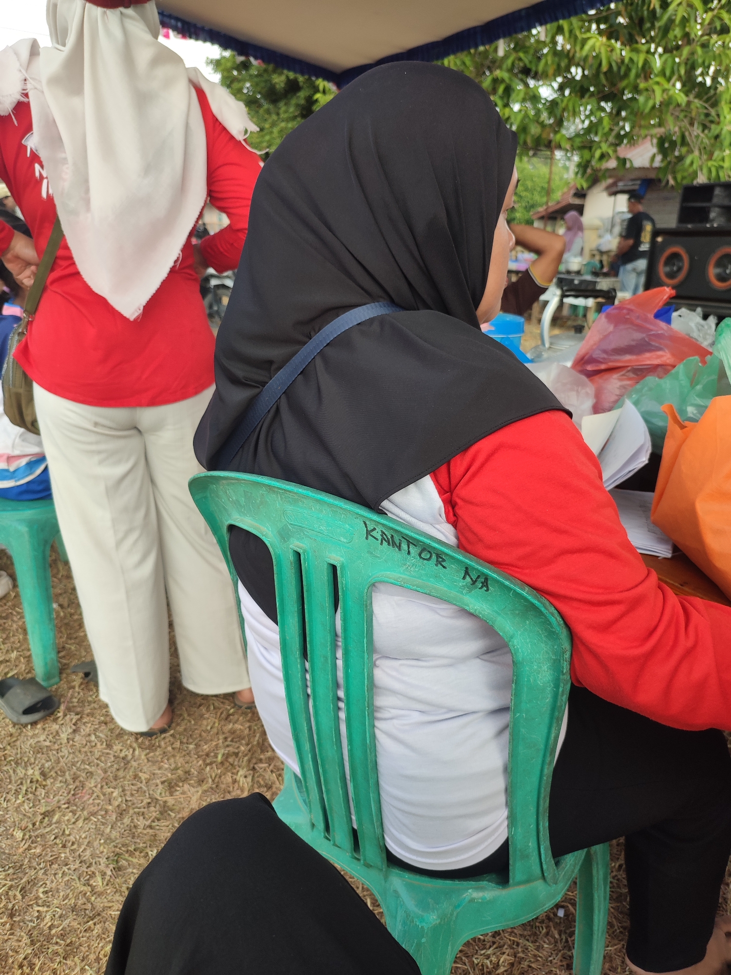 The image shows a rear view of a person wearing a black hijab and a red shirt, sitting on a green plastic chair. The chair has the text "KANTOR NA" written on its backrest. To the left of the person, another individual is partially visible, wearing a white hijab and a red shirt, and light-colored pants. In the background, there is a tent or canopy, trees, and a building. Several plastic bags, including orange and red ones, are scattered on a table in the background, along with papers. A speaker system is also visible in the background. The ground appears to be covered in dry grass and dirt. The scene suggests an outdoor gathering or event. The location is identified as Nusa Jaya, Indonesia.