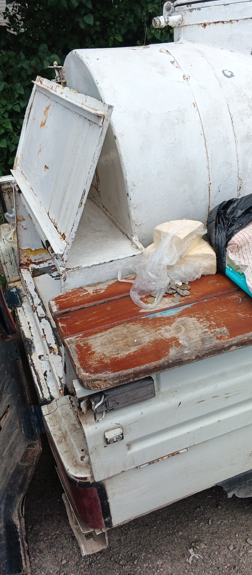 The image shows the rear of a white, weathered, and rusty water truck. The truck bed is wooden with visible wear and tear. On the truck bed, there are two rectangular blocks wrapped in plastic sheeting, a pile of coins, a black plastic bag, and a folded patterned cloth. The background consists of green foliage. The scene appears to be in an outdoor, possibly rural or industrial, setting. The specific location in Smila, Ukraine, is not determinable from the image alone.