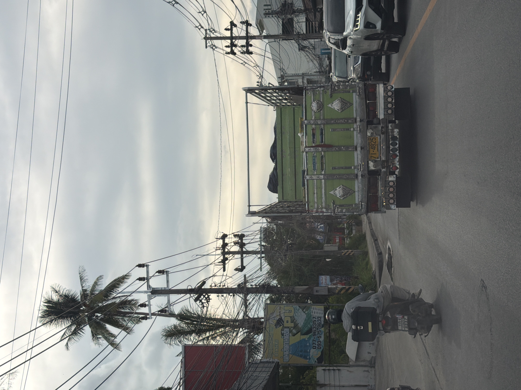 The image depicts a street scene in Baan Bo Phut, Thailand, likely during the daytime given the ambient light. The perspective is from a vehicle traveling on the road, as evidenced by the road surface in the foreground and the rear of vehicles ahead.
The primary subjects include a large green truck with a metal frame on its back, and a person riding a motorcycle. The truck has intricate decorative elements on its sides and a license plate visible with the text "71-4825". The motorcycle rider is wearing a grey hooded jacket and a helmet, and is seen from the side and rear, with a black storage box mounted on the back of the motorcycle. The motorcycle also has a visible license plate.
The setting is a busy street with numerous power lines crisscrossing overhead, supported by utility poles. A palm tree is visible on the left side of the frame, with its fronds extending into the sky, suggesting a tropical location. Buildings and other vehicles are present in the background, contributing to the urban street environment. There are also some advertisements and signs visible, though the text is largely unreadable or partially obscured. One sign appears to have text that might be in Thai.
The weather appears to be overcast, with a diffused sky that is light gray, indicating possibly a cloudy day or approaching rain. There are no strong shadows, which supports this observation. The scene conveys a sense of everyday movement and activity on a street in Thailand.