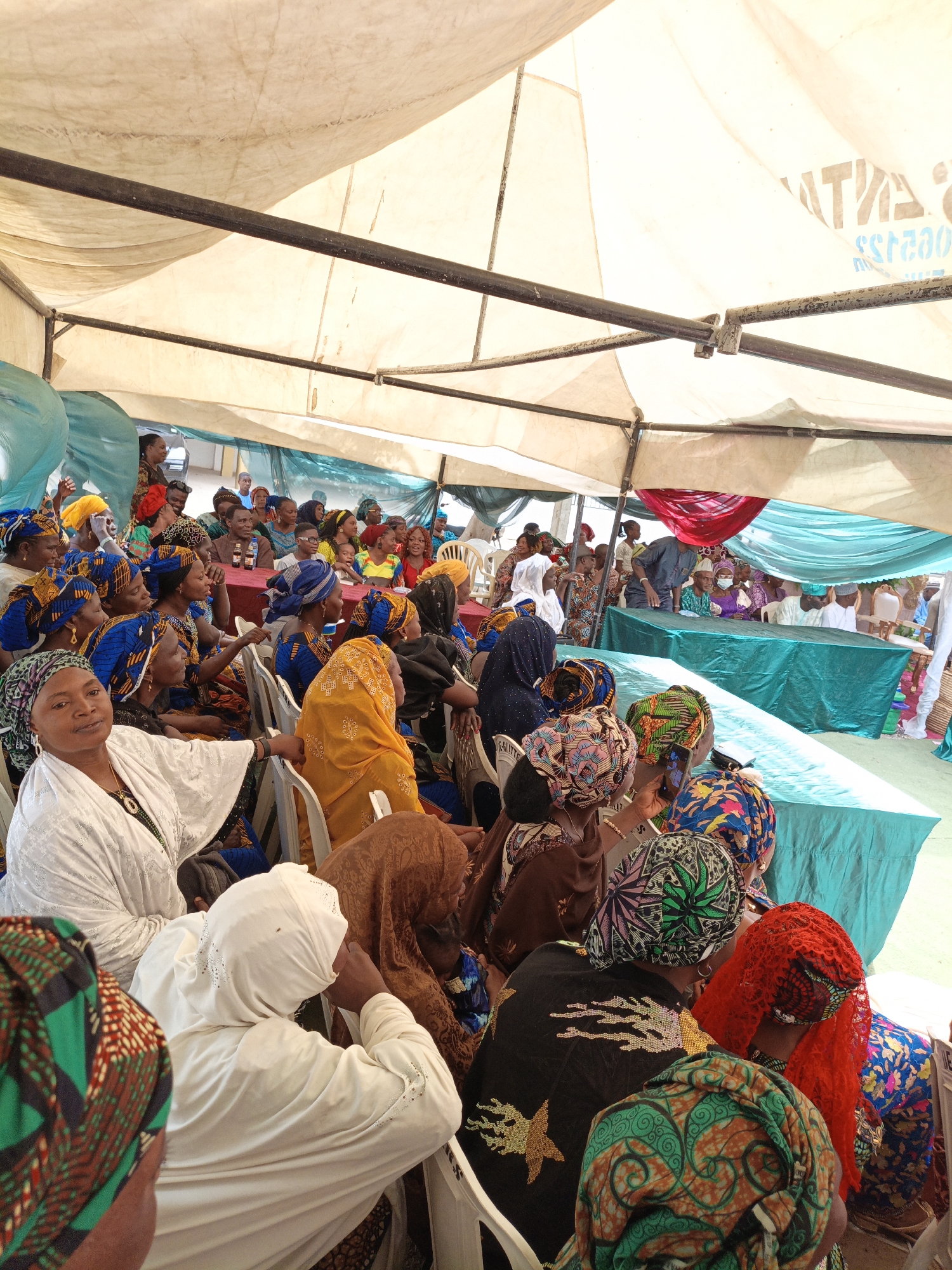 This image captures a vibrant community gathering under a large tent, likely in Sabon Gari West, Nigeria. The scene is densely populated with people, primarily women, seated on white plastic chairs. Many attendees are adorned in colorful traditional African attire, including brightly patterned fabrics and elaborate head wraps (gele) in hues of blue, yellow, green, red, and brown. A few men are also visible, particularly towards the rear and at the front of the seating arrangement.
The event takes place under a light-colored canvas tent supported by black metal poles, providing shade from the daytime sun. Tables covered with teal or turquoise tablecloths are arranged at the front, suggesting a head table or a speakers' platform. One table in the mid-ground holds what appears to be a bottled soft drink. Decorative teal fabric drapes and a splash of red fabric are also visible inside the tent.
The attendees appear engaged, with many looking towards the front of the gathering. One woman in the immediate foreground, wearing a white outfit and head covering, looks directly at the camera with a calm expression. The bright, diffused lighting indicates it is daytime. The ground beneath the tent seems to be bare earth. Faint, mirrored text, possibly "NATAS SERVICES" or similar, is visible on the tent fabric in the upper right background. The gathering has a communal and attentive atmosphere.