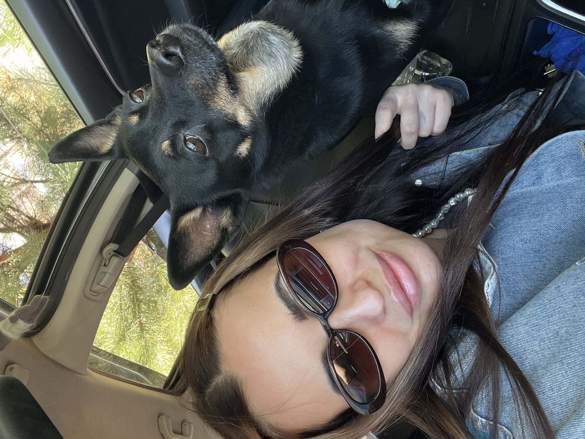 A woman wearing sunglasses and a denim jacket is taking a selfie inside a car with a black and tan dog lying upside down next to her. The car is parked near trees, which are visible through the window. The location appears to be Unknown, Unknown.