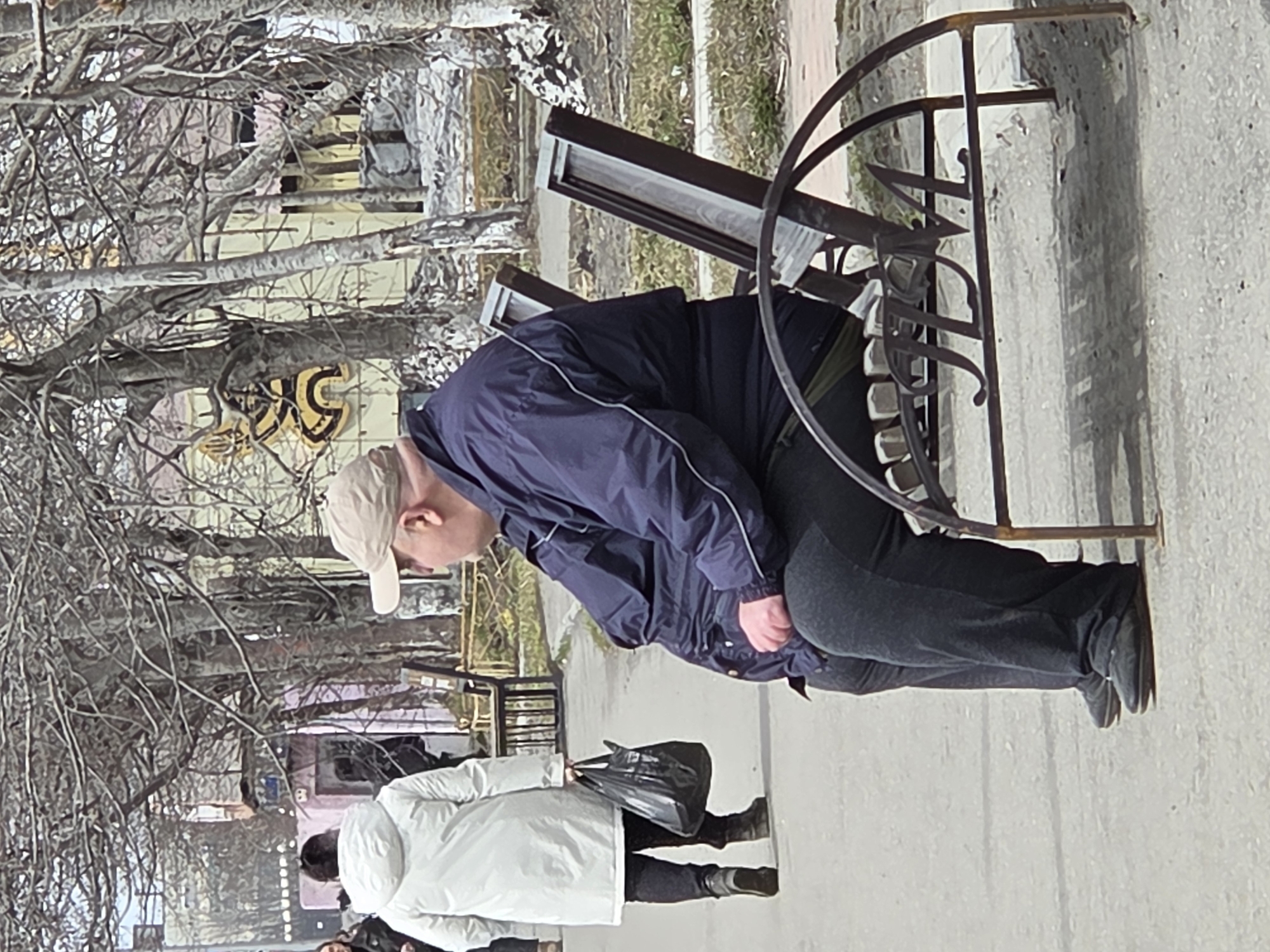 A pedestrian scene on a city street in Unknown, Unknown. A person wearing a navy jacket, grey pants, black shoes, and a beige baseball cap is bending at the waist looking at a small metal fence with a decorative circular design at the top. The person is standing on a light grey sidewalk. Behind them and to the left is another person wearing a white coat, black pants, and black shoes, holding a black bag. In the background, there are bare trees, buildings with graffiti, and hints of other people in the distance. The overall light is muted and diffused, giving the image a slightly overcast feel.