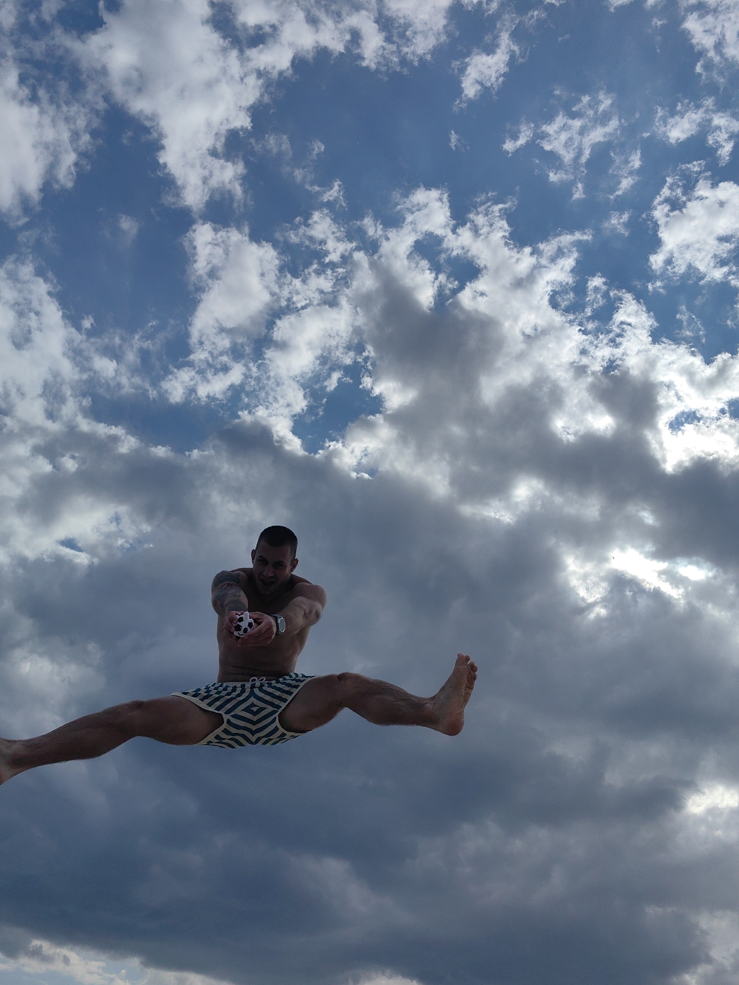 A man is captured mid-air performing a split jump against a backdrop of a dramatic, cloudy sky, likely above a beach in Varna, Bulgaria. The man, who appears to be in athletic condition, wears striped swim trunks. He is holding a small, spherical object in his hands, which is black and white and resembles a miniature soccer ball. His expression is one of exertion and perhaps joy as he reaches for the object. His tattoos are visible on his arms. The lighting suggests it is daytime, with the sun breaking through some of the darker clouds. The perspective is from below, emphasizing the height and dynamism of the jump against the expansive sky.