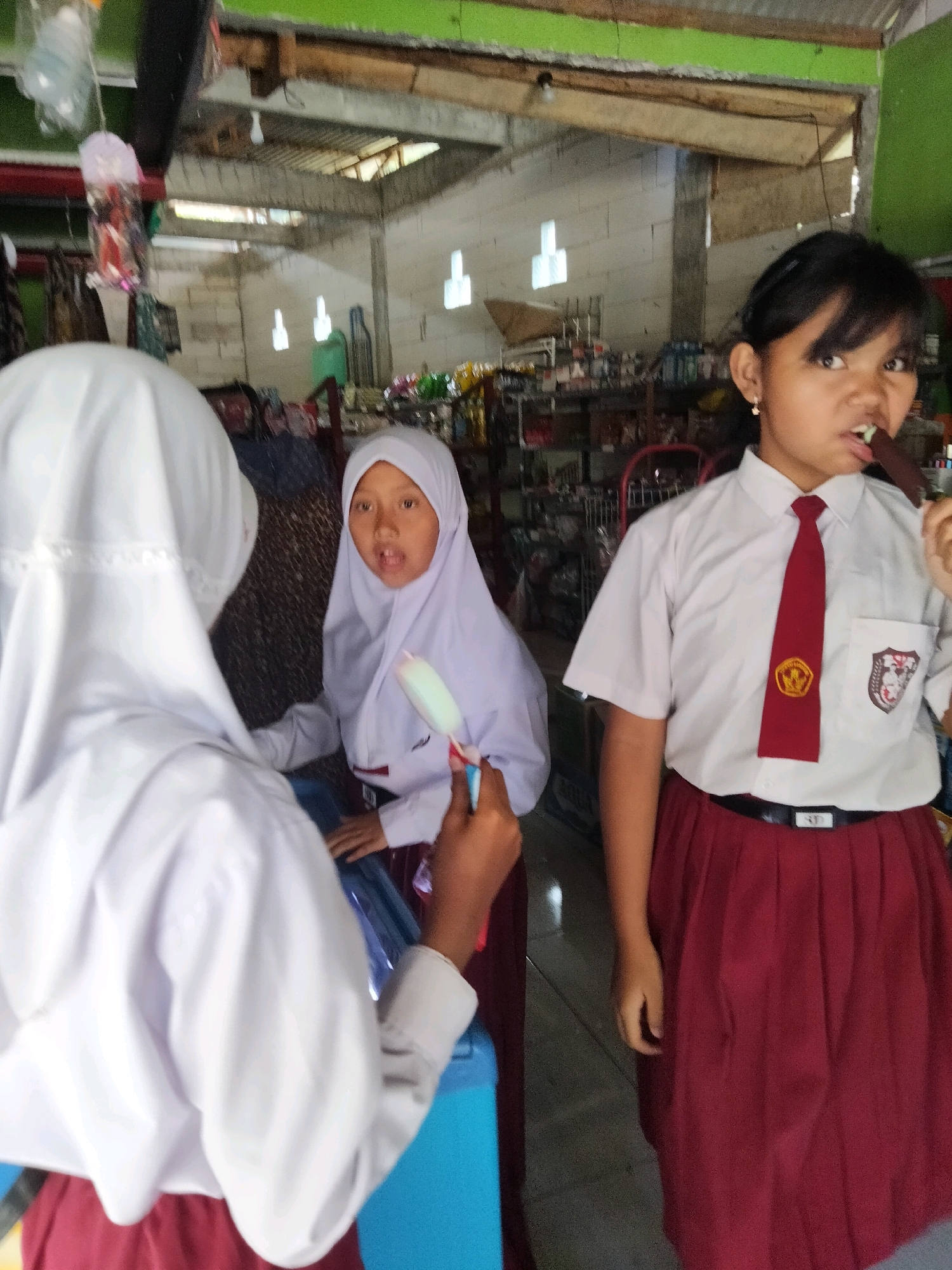 In a market stall in Sidorejo, Indonesia, three young girls in school uniforms are pictured. The girl on the right, facing the camera, is wearing a white shirt with a red tie and a maroon skirt. She is holding a dark-colored ice cream bar to her mouth. The girl in the middle, also in a white uniform and hijab, is looking to her left and appears to be holding a light-colored ice pop. The third girl, on the left, is mostly obscured from view, with only her white hijab and a portion of her white shirt and maroon skirt visible. She is facing away from the camera and towards the girl in the middle. The background suggests an indoor market setting with shelves stocked with goods and a partially exposed ceiling.