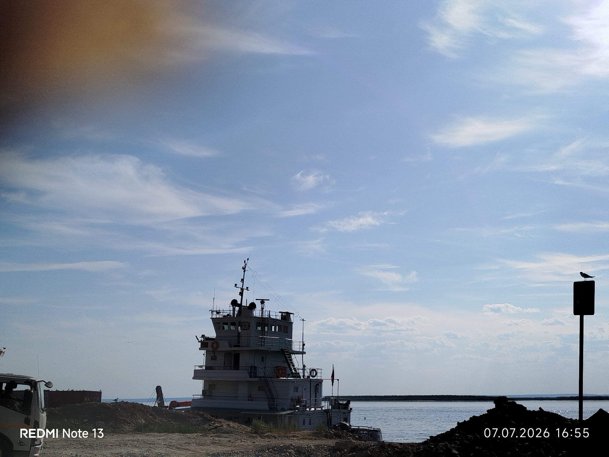 The image depicts a waterfront scene in Nizhny Bestyakh, Russia, on July 7, 2026, at 16:55. A large, multi-story white tugboat is moored near the shore. In the foreground, there is a pile of dark soil and rocks. To the left of the tugboat, the front of a white truck is visible, along with a dark, flat-topped structure. The water extends to the horizon, meeting a clear blue sky with scattered white clouds. A dark post with a bird perched on top stands to the right of the frame. The lower-left corner displays "REDMI Note 13" and the lower-right corner shows the date and time.