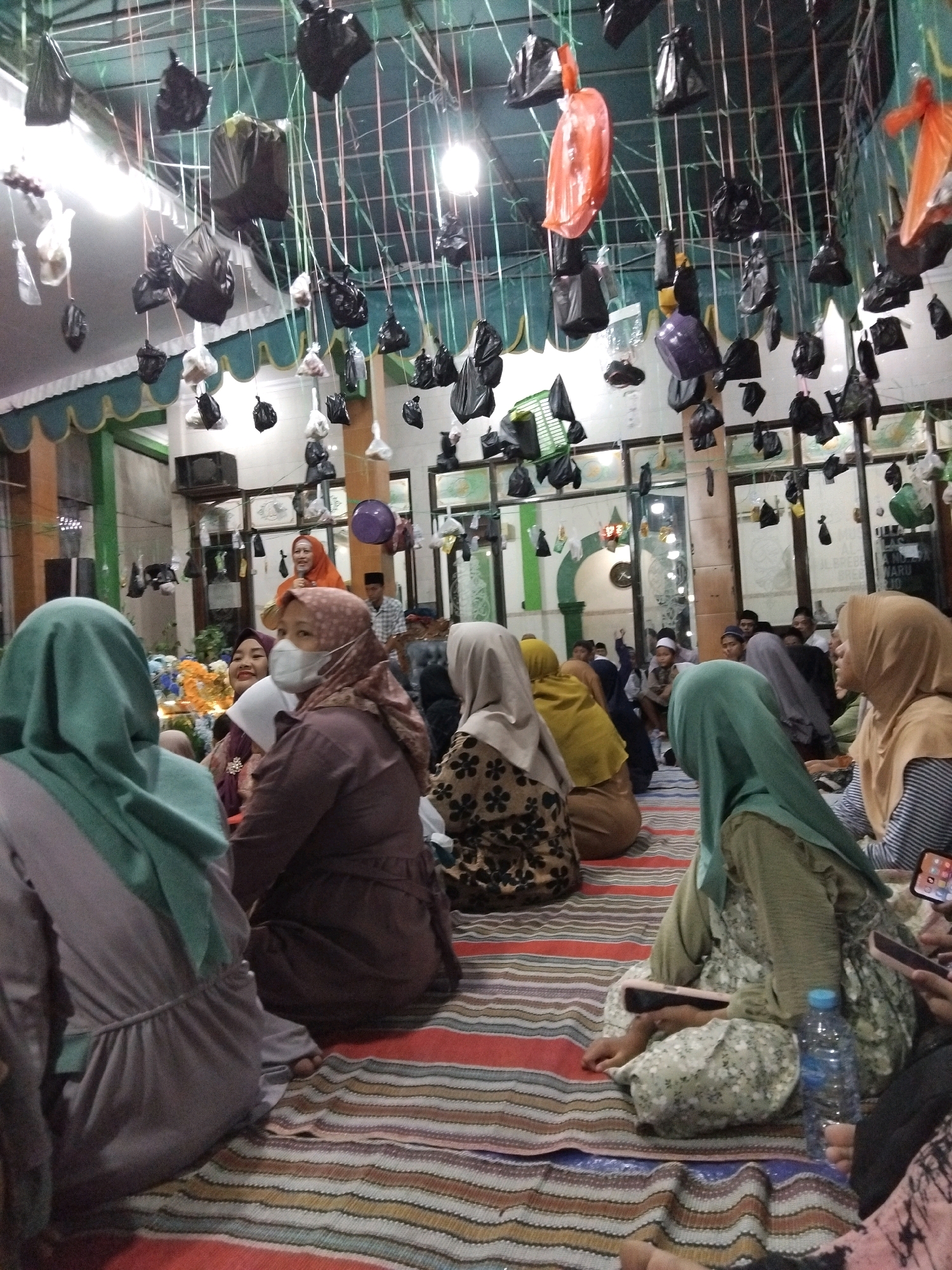 In Kepuh Kiriman, Indonesia, a group of people, predominantly women in hijabs, are gathered indoors. The room appears to be a communal space, possibly a religious building, with a low ceiling from which numerous black plastic bags and some orange and purple plastic items are suspended by strings. The floor is covered with a striped mat. Several individuals are seated on the mat, some with their backs to the camera, while others are facing it. One woman in the foreground wears a light brown hijab and a mask. Another woman to her left is in a grey-green hijab. A woman in the background, wearing an orange hijab, is smiling and looking upwards. The environment suggests a gathering or event, with the suspended bags potentially part of a decorative or ceremonial display.