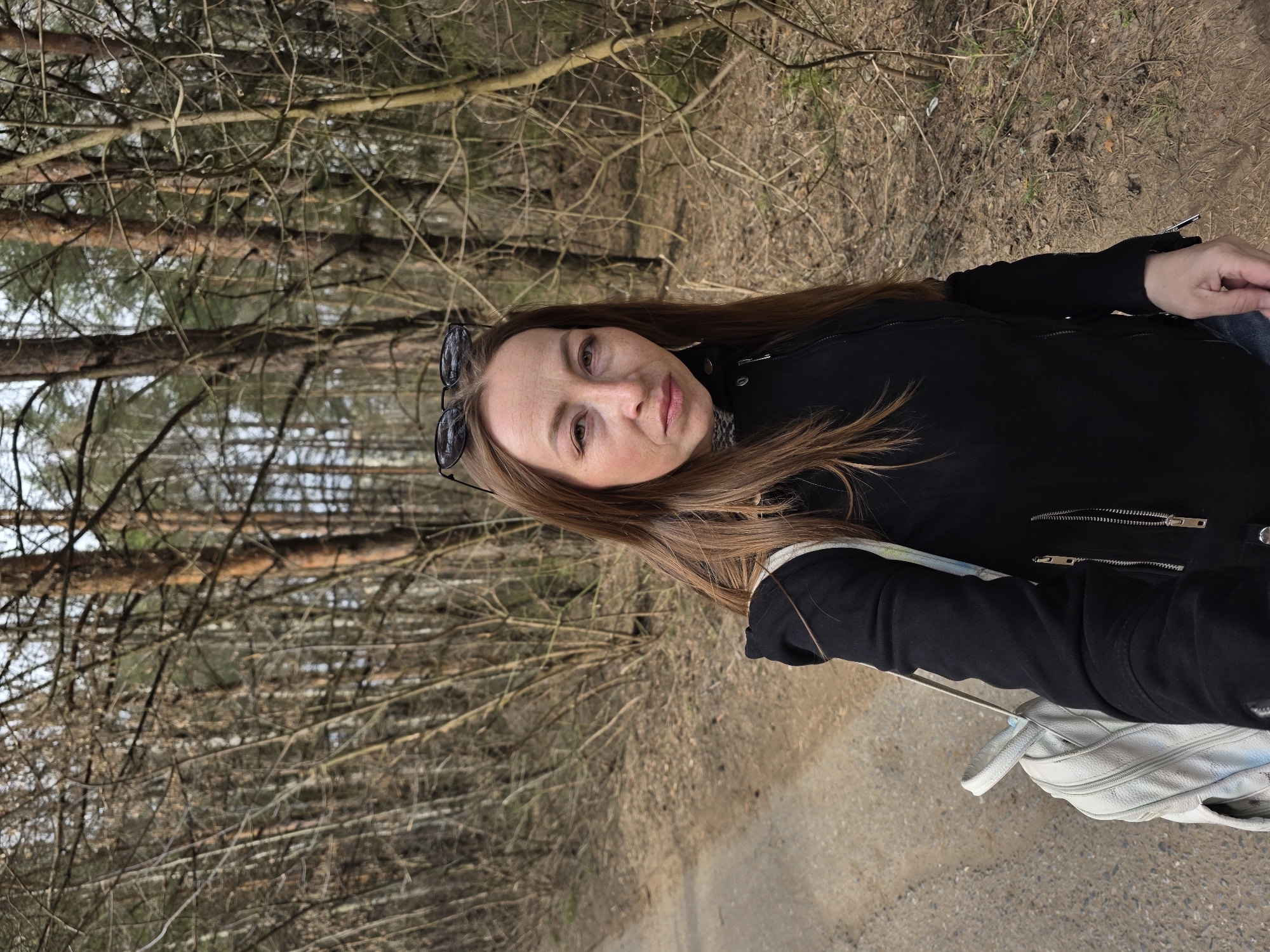 A woman with long brown hair is positioned in the foreground of the image. She is wearing a black jacket and has sunglasses perched on her head. A white backpack is visible on the ground next to her. The background consists of a dense forest with numerous trees. The ground is covered in dry leaves and branches. Based on the natural environment, it is plausible this image was taken in a wooded area in or near Chaikovsky, Russia.