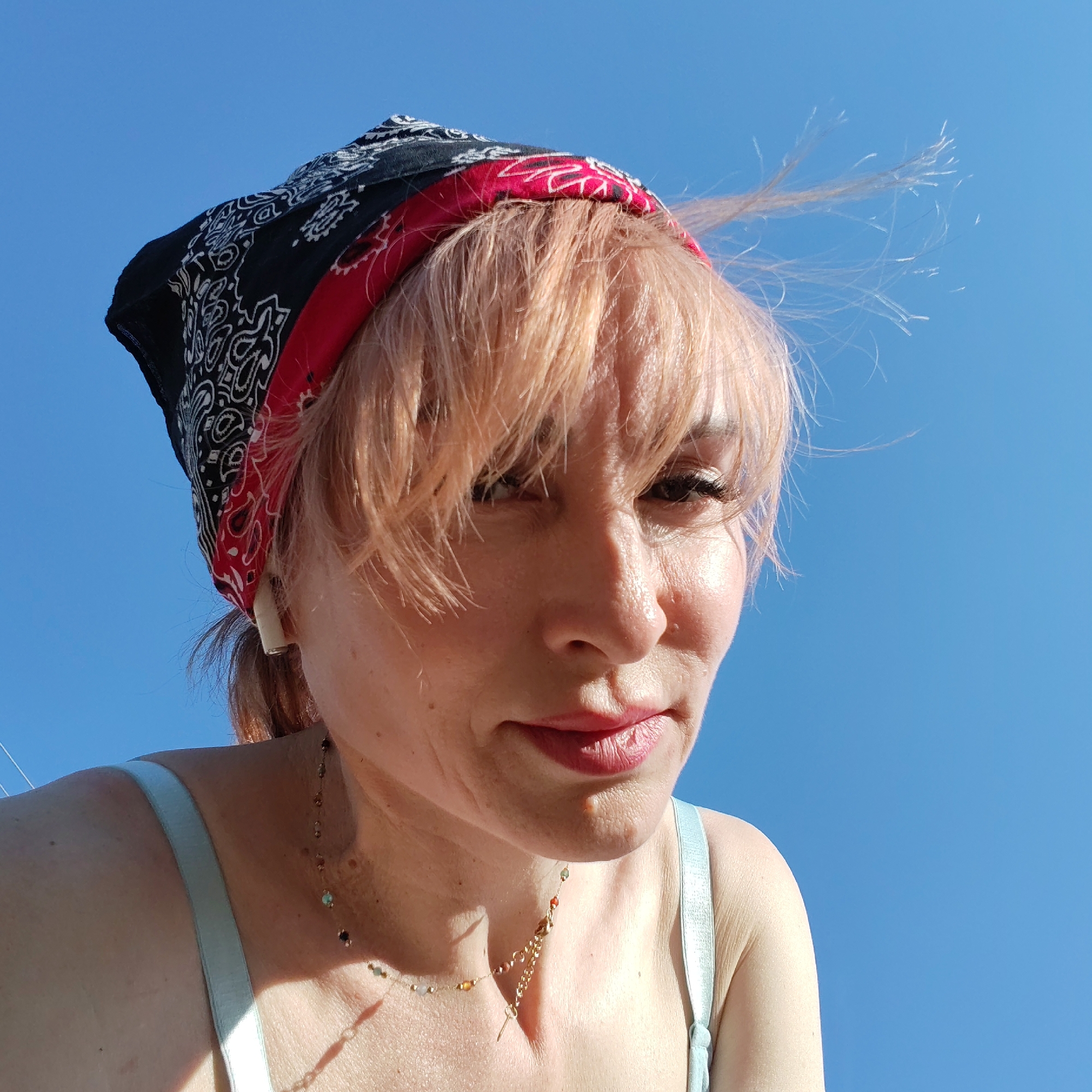 A woman with light pinkish-blonde hair wearing a black and red paisley-patterned bandana is seen from a low angle against a clear blue sky. Her hair is somewhat windblown, with strands lifting off her forehead. She has fair skin with visible pores and some freckles. She is wearing a light blue tank top and a delicate necklace with small colored beads and a key pendant. Her lips are painted with a rose-colored lipstick, and her expression is slightly pensive. The location is not specified in the image and the city of Chaikovsky, Russia is not discernible from the image alone.