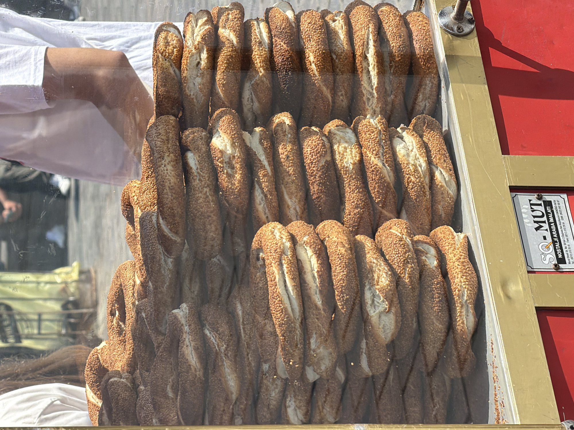 A display case filled with stacked simit, a Turkish bread, is positioned in front of a vendor selling them in the Fatih district of Istanbul, Turkey. The simit are round and covered in sesame seeds, with some appearing darker and crispier than others. Behind the display case, a person wearing a white t-shirt and jeans is visible, with their right arm slightly bent as if serving or preparing the simit. The background shows a blurred street scene with people and buildings. A sign on the front of the display case provides vendor information, including "SQ - MUT NOSTALJİ ARABA İMALATI" and contact details. The overall scene suggests a busy street food vendor.