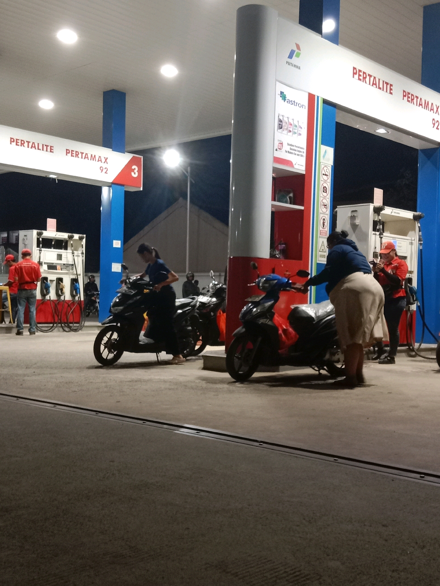 The image captures a nighttime scene at a Pertamina gas station in South Tangerang, Indonesia, likely on Jalan Pondok Indah. In the foreground, a woman in a dark top and a beige skirt is standing next to a dark-colored scooter. To her left, another scooter is parked, and a woman in a dark blue shirt and dark pants is seated on it.
Further back, a Pertamina gas pump is visible, with two attendants in red uniforms standing nearby. One attendant is operating the pump while the other, wearing a red cap, is looking at their phone. Several other motorcycles are parked in the background. The gas station canopy is illuminated by overhead lights, and signage indicating "PERTALITE" and "PERTAMAX 92" is prominent. The overall environment appears to be a standard gas station forecourt.