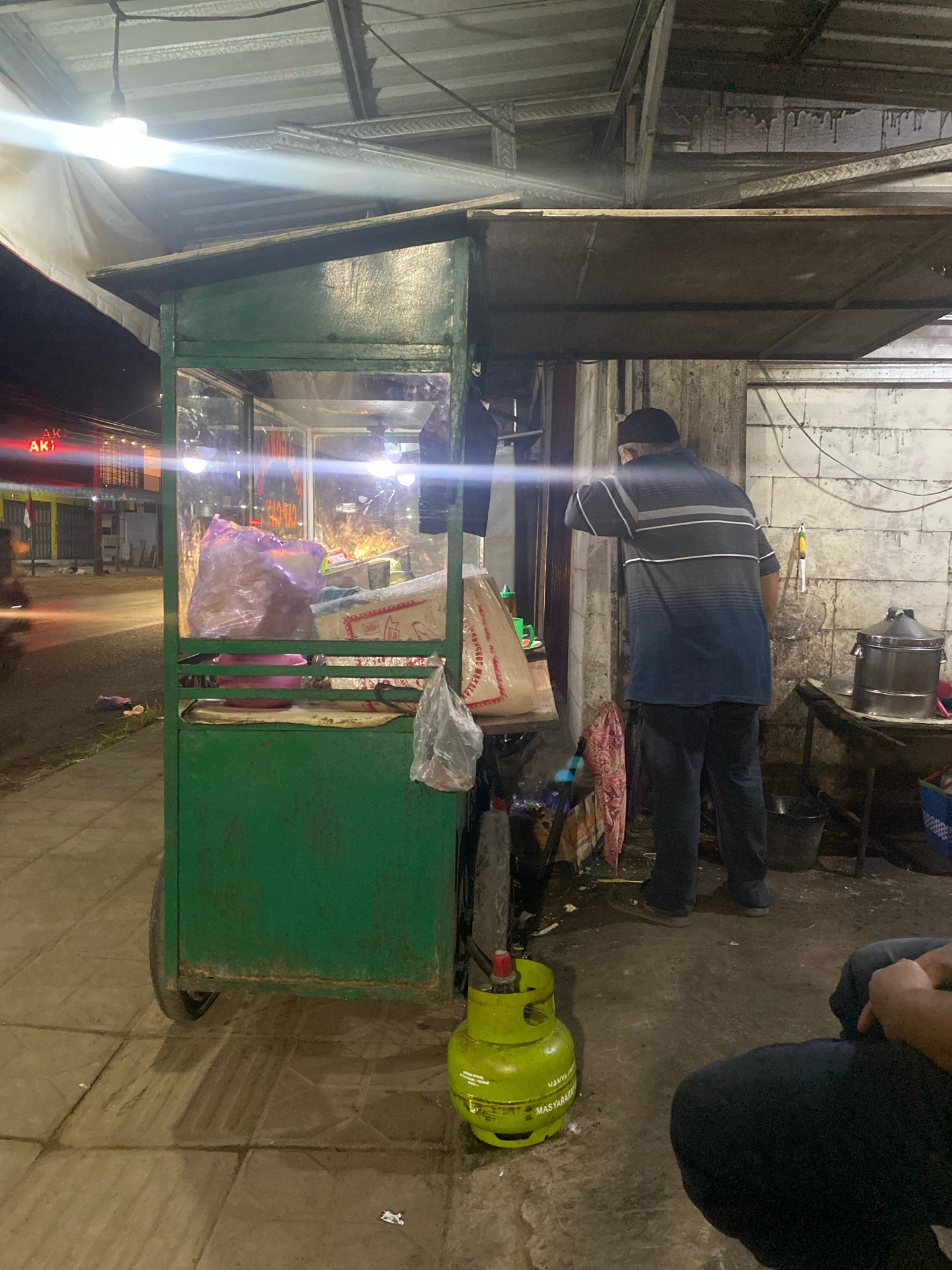 In Pegaden Tengah, Indonesia, a man wearing a dark skullcap and a grey and blue striped polo shirt stands with his back to the camera, leaning towards a green food cart. The cart, which has glass panels displaying various items within, is positioned on a paved sidewalk. A bright green gas cylinder with visible text is located in the foreground, to the right of the food cart. To the right of the man, a metal table holds a large stainless steel pot with a lid, and a bucket is on the ground. Further to the right, a portion of another person is visible, seated and wearing dark clothing. The environment appears to be an outdoor commercial area at night, with artificial lighting illuminating the scene. The background shows a street with buildings and closed storefronts, suggesting a business district in the city.