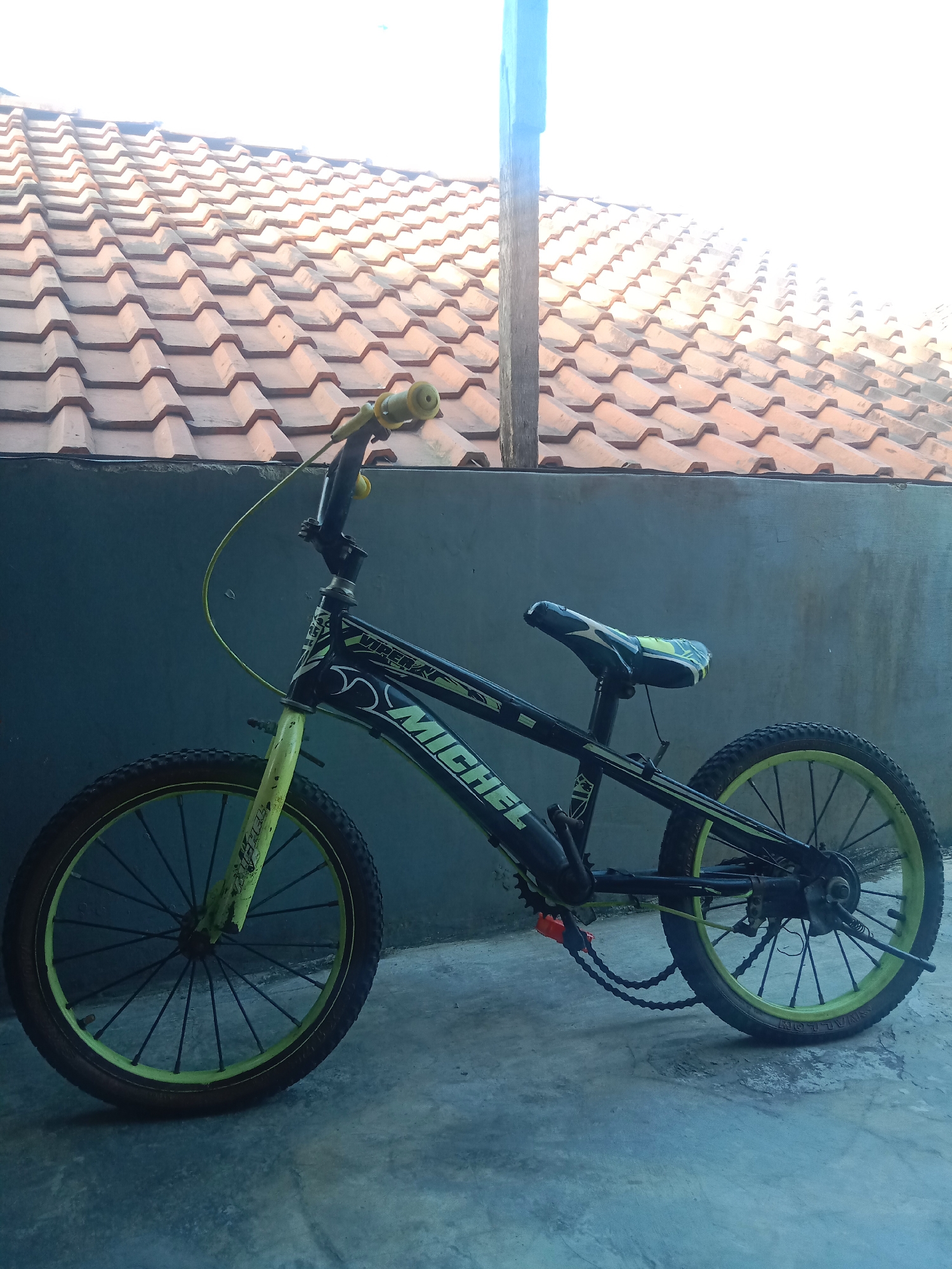 The image displays a BMX-style bicycle parked on a concrete surface in what appears to be an outdoor area. The bicycle is black and yellow, with a seat and handlebar grips. It is positioned against a textured gray wall, with a tiled roof visible in the background. The sunlight suggests it is daytime. The presence of the bicycle and the tiled roof suggest a residential setting, possibly a backyard or alleyway. Kutosari, Indonesia is a village in Central Java, known for its agricultural landscape and traditional houses often featuring tiled roofs.