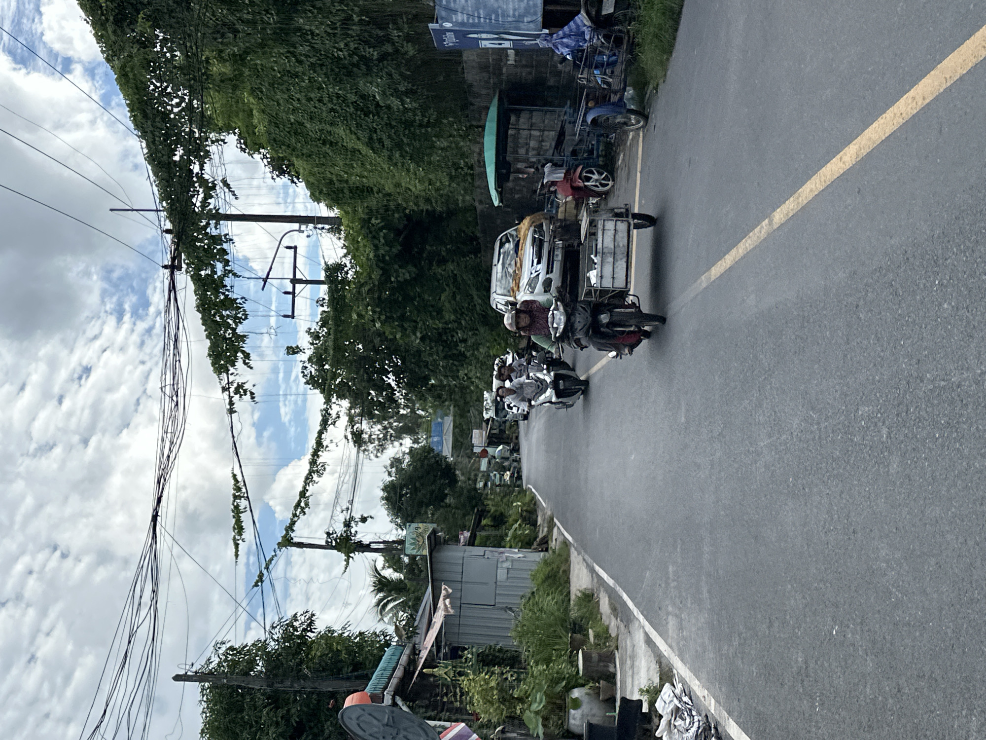 A street scene in an urban environment shows a paved road with a yellow line down the center, bordered by vegetation and structures on either side. Several motorcycles are present, some with sidecars, and a white SUV is parked on the right side of the road. The sky is partly cloudy. Power lines are visible overhead, supported by wooden poles. A person wearing a helmet is riding a motorcycle with a sidecar loaded with items, moving along the road. In the background, other vehicles and structures are visible.