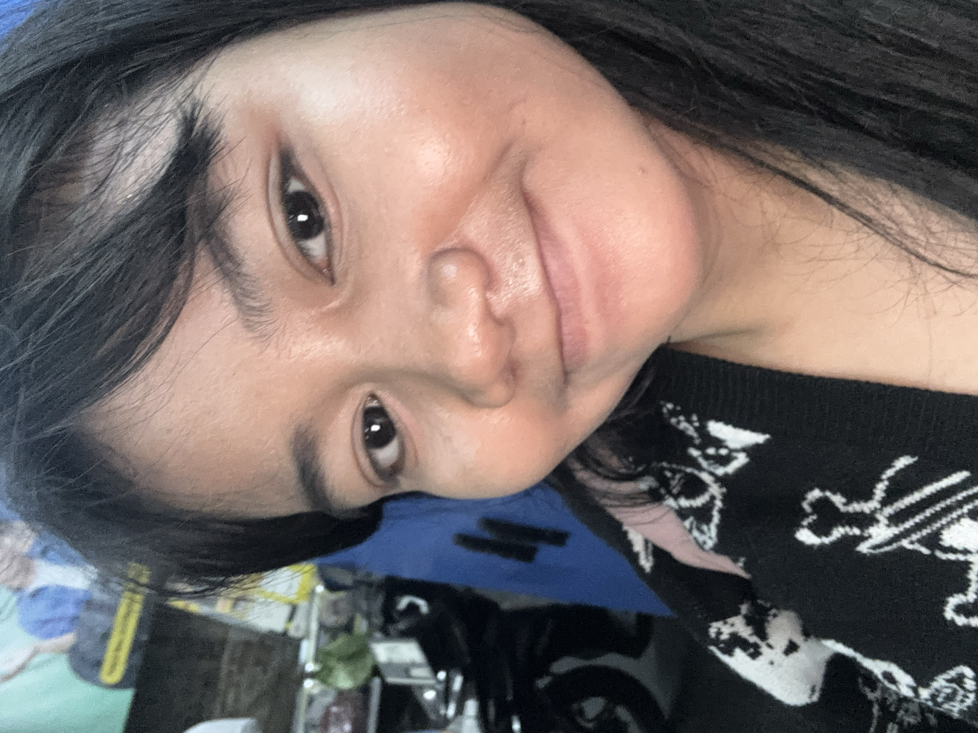 A close-up, eye-level, indoor, selfie shows a young woman with dark hair and a black and white patterned sweater. The woman is looking directly at the camera with a slight smile. Her skin appears slightly oily, and there is some faint makeup visible on her face. Her hair is long and straight, falling around her face and shoulders. The background is blurred, but appears to be an indoor setting with some objects and a blue curtain visible. The overall lighting is warm and natural. The image was taken in North Jakarta, Indonesia.