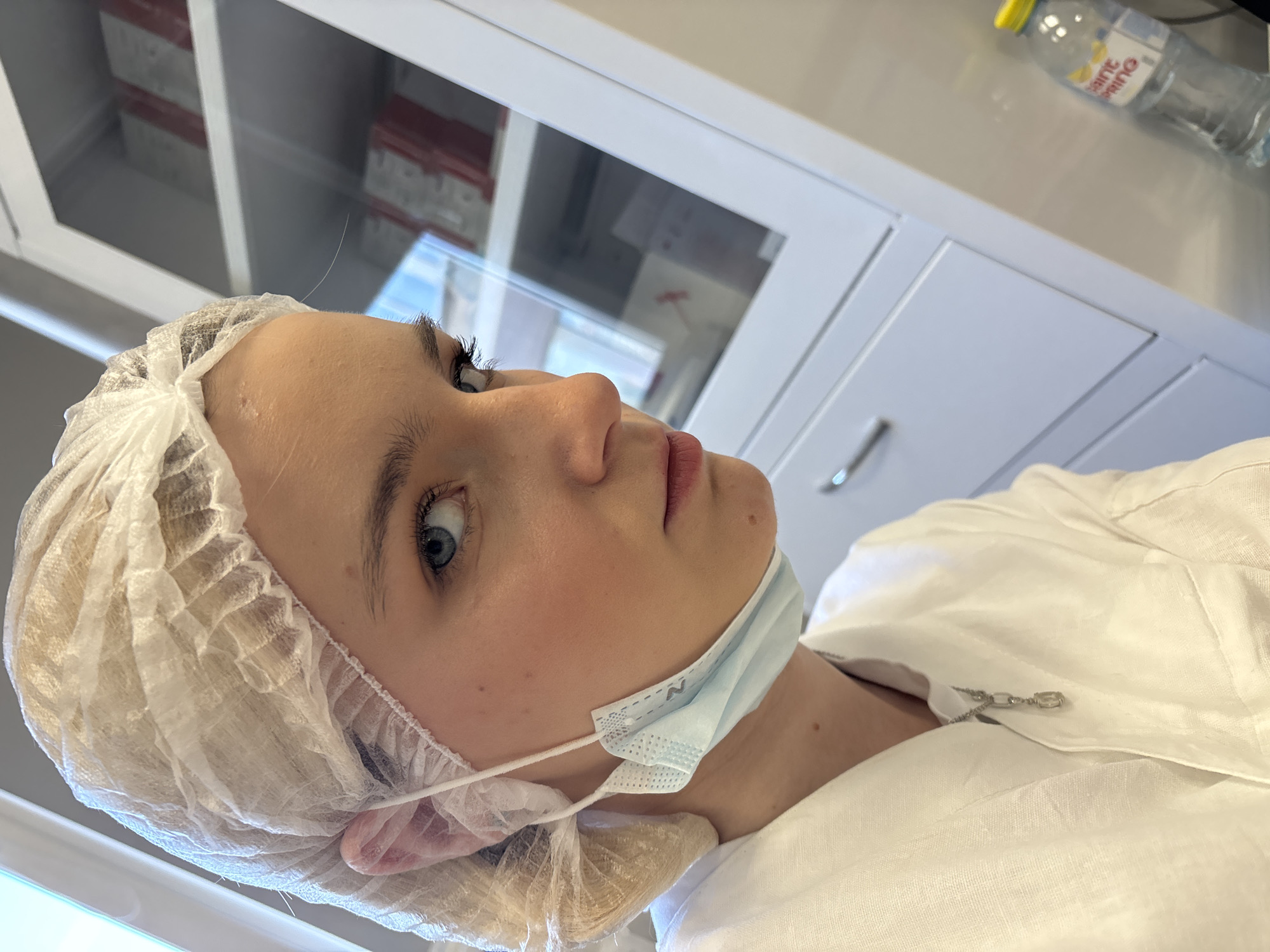 A young woman wearing a disposable hair net and a light blue surgical mask is the primary subject of the image. The woman's skin is fair, and her hair appears light blonde beneath the net. The mask covers her mouth and nose, secured by white elastic strings. She is also wearing a white garment, possibly a lab coat or medical scrubs, fastened with a small decorative clasp.
The background suggests a clinical or medical setting. A white cabinet with glass-fronted doors is partially visible, revealing stacked items within, possibly medical supplies or boxes. To the right, a white countertop is visible with a bottle of water.
The photograph's perspective is slightly angled, with the camera positioned above and to the side of the subject.
Due to the clinical setting and the person's attire, it can be assumed the location is somewhere in Saint Petersburg, Russia.