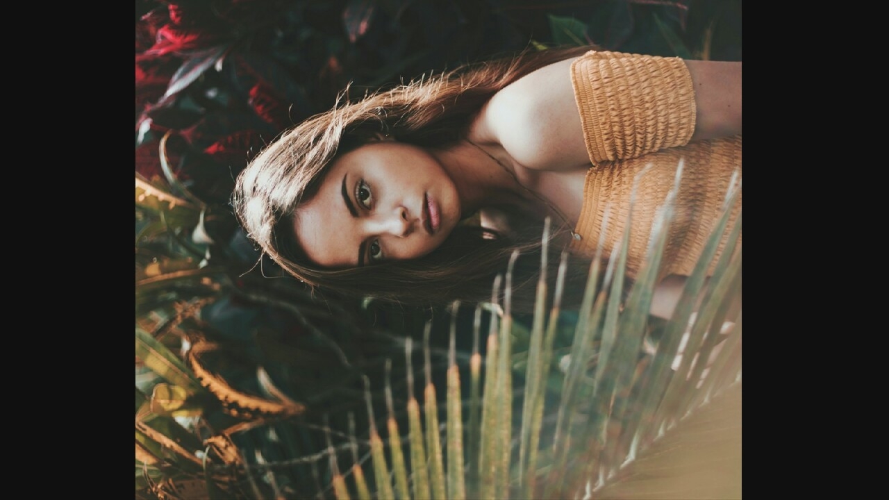 A portrait of a young woman with long, brown hair is captured in an outdoor setting. She is wearing an off-the-shoulder, ruffled yellow top and a thin necklace. The background is filled with lush, green foliage, with hints of red and yellow in the leaves, suggesting a tropical or garden environment. A frond from a palm tree is prominently placed in the foreground, slightly blurring the left side of the image. The woman is looking directly at the camera with a neutral expression. The lighting is soft, casting gentle shadows on her face.